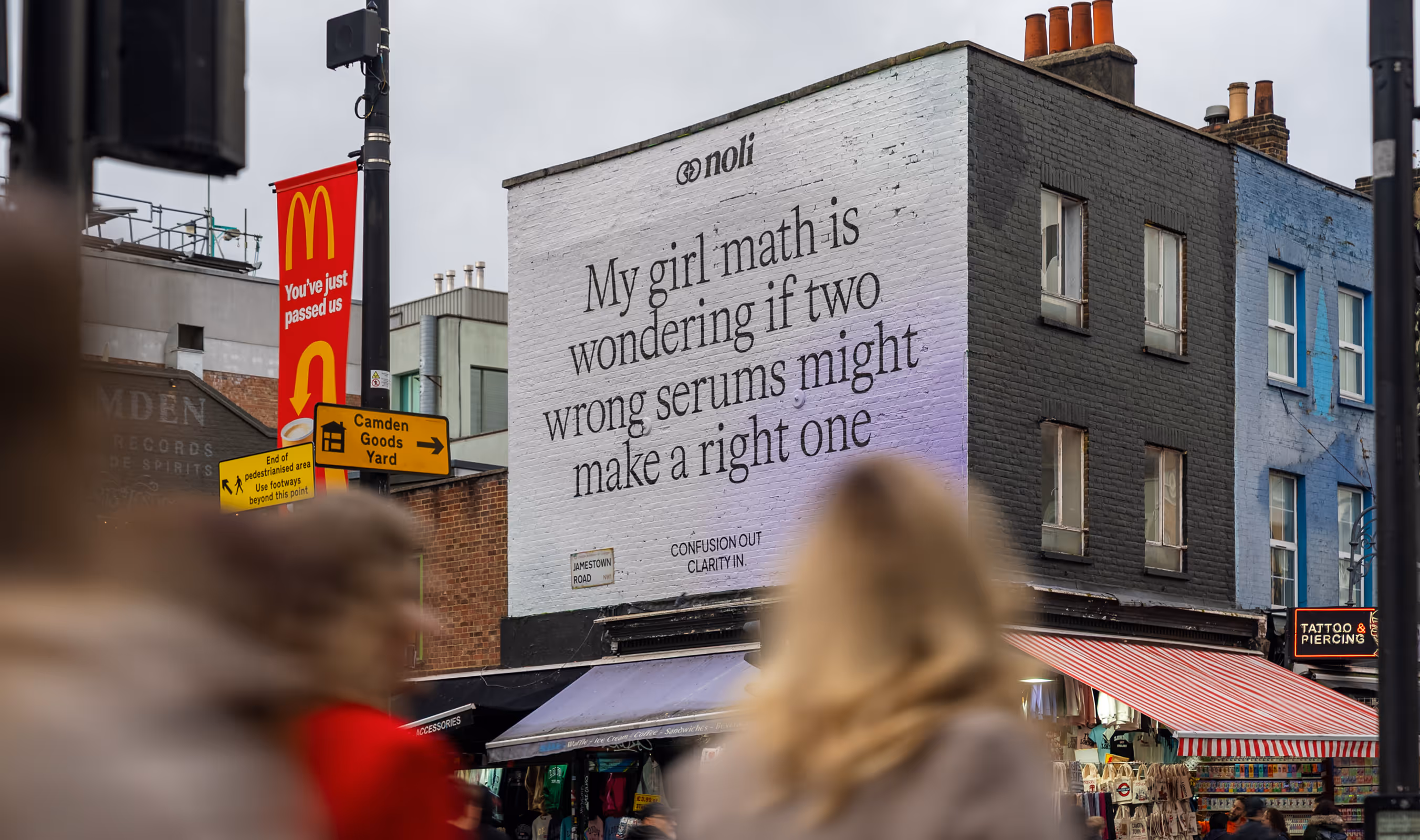 City street scene with a white brick wall displaying a large text mural reading 'My girl math is wondering if two wrong serums might make a right one', surrounded by shops and pedestrians.