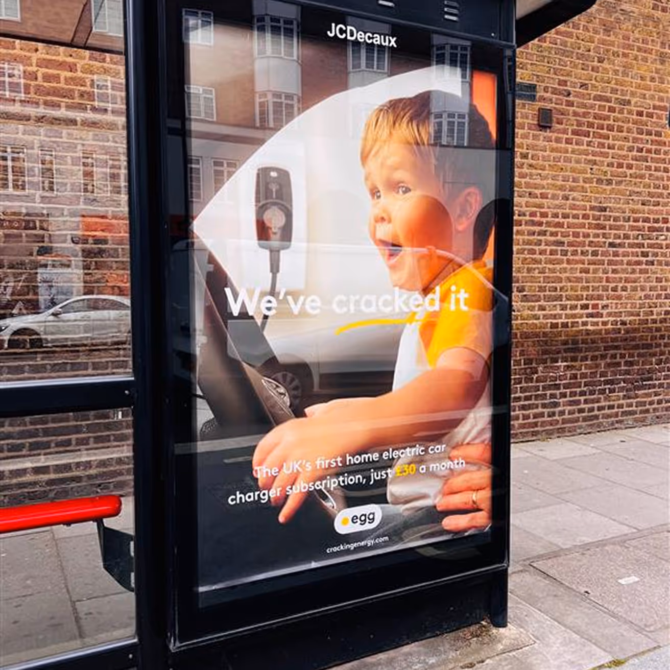 Bus stop advertisement featuring a smiling toddler holding a steering wheel with text about the UK's first home electric car charger subscription for £30 a month.