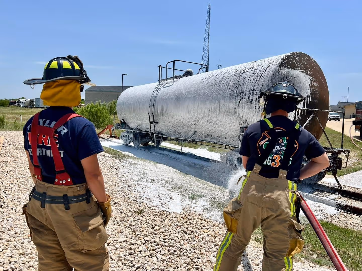 Two firefighters in protective gear spraying foam on a large cylindrical tank outdoors.