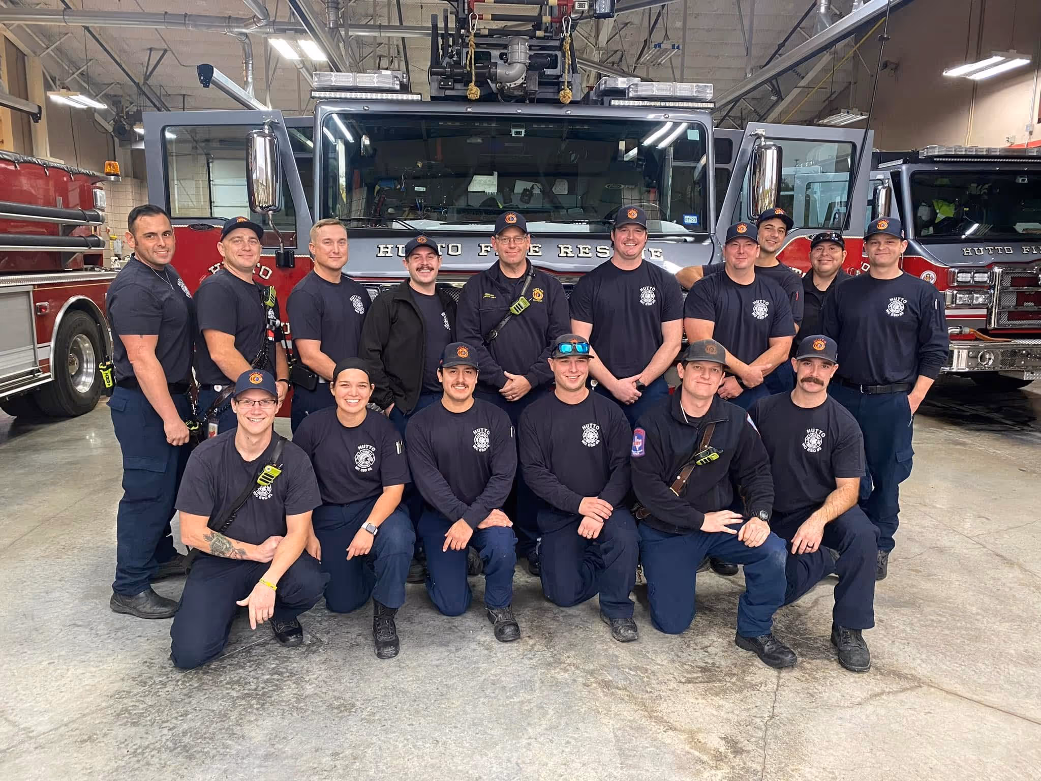 Group of firefighters in uniform posing in front of fire trucks inside a fire station.