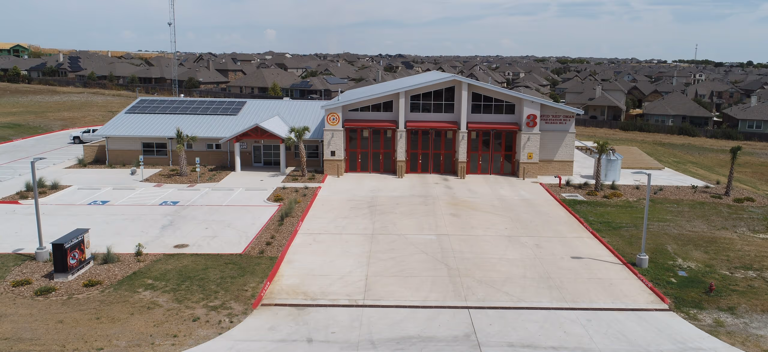 Fire station building with three red garage doors, solar panels on the roof, and parking lot with handicap spaces.