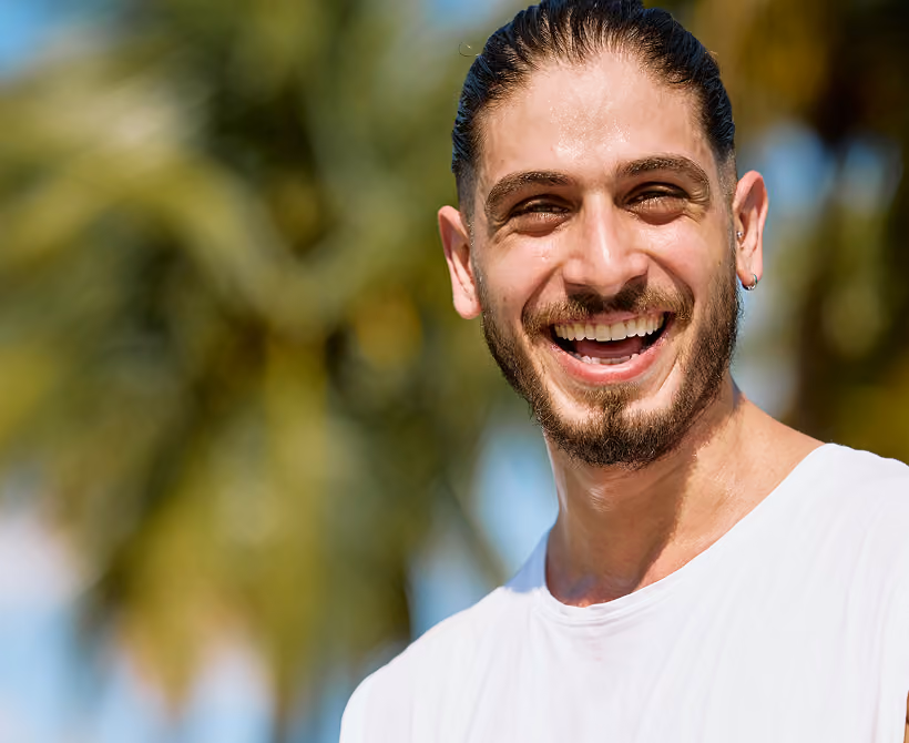 Smiling man with wet hair and beard in white shirt, palm trees in blurred background.