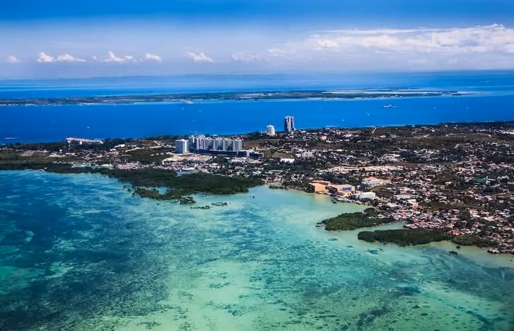 Aerial view of coastal city with clear turquoise waters meeting urban buildings under a partly cloudy sky.
