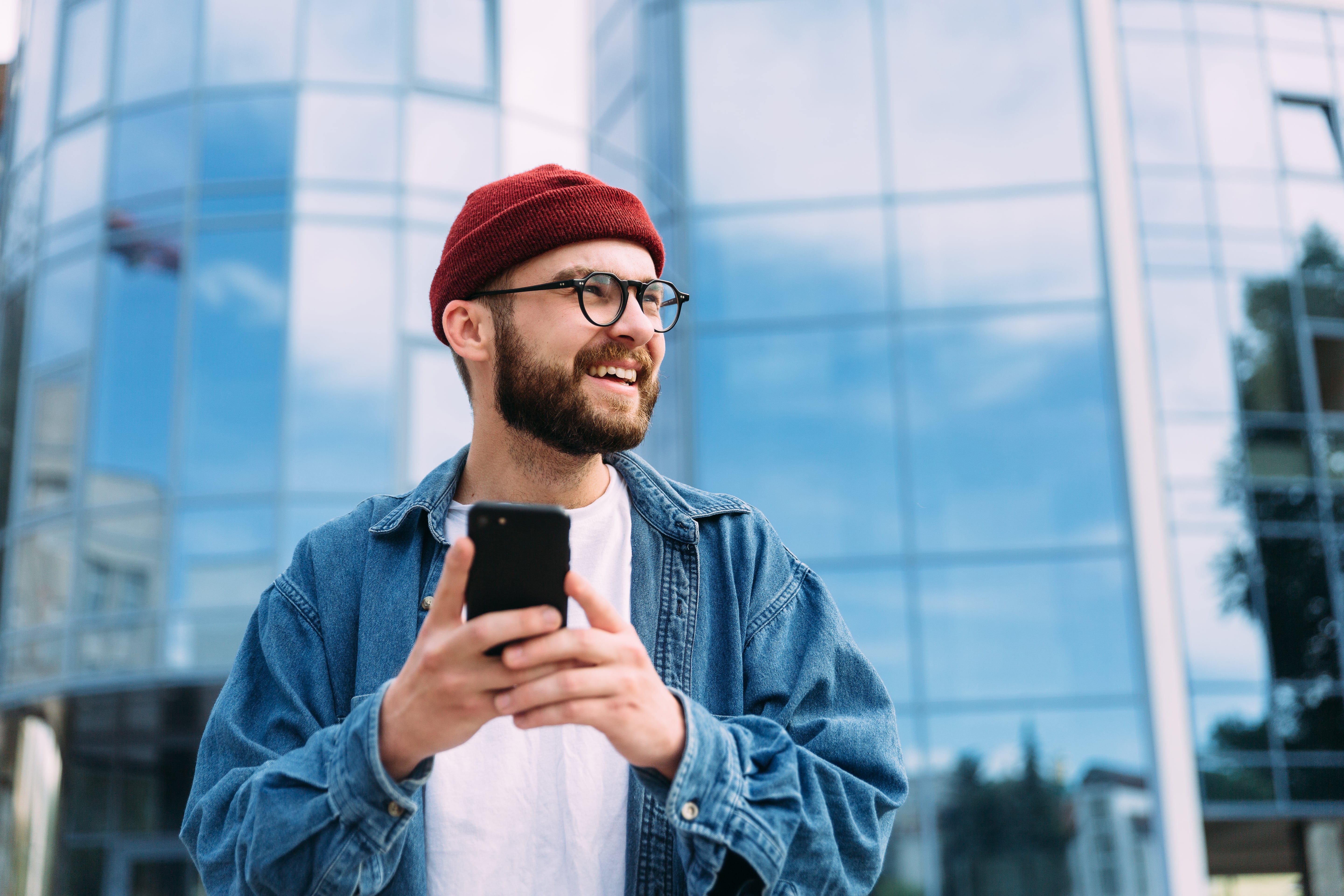 Smiling bearded man wearing glasses and a red beanie holding a smartphone with a glass building in the background.