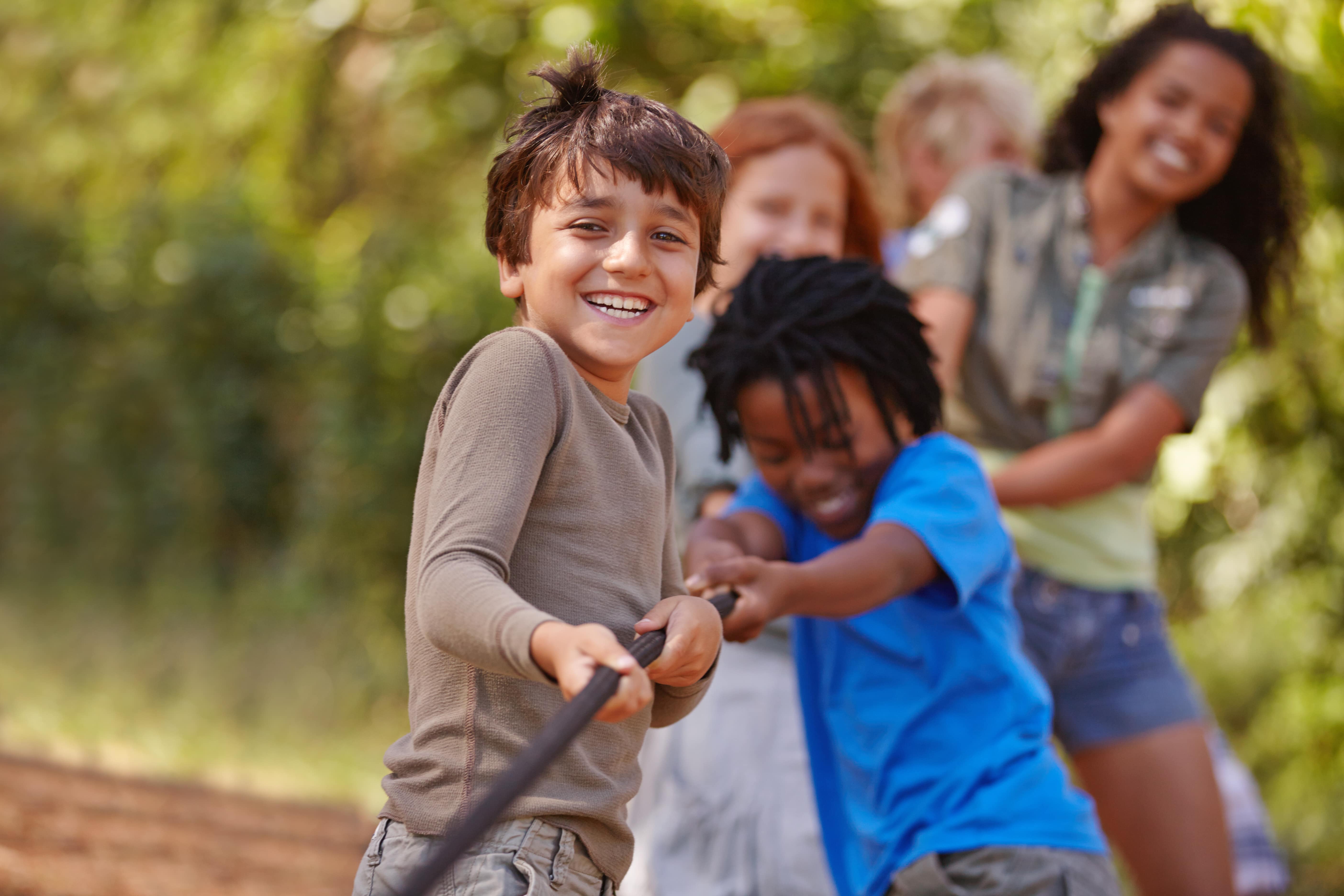 A group of diverse children playing tug-of-war outdoors on a sunny day, with one boy smiling at the camera.