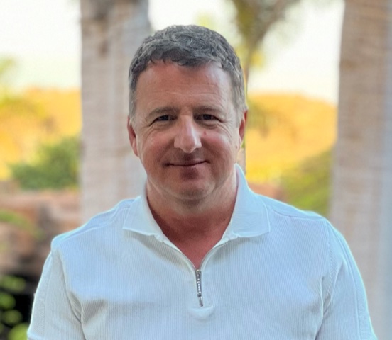Middle-aged man with short gray hair wearing a white quarter-zip shirt, standing outdoors with blurred trees and columns in the background.