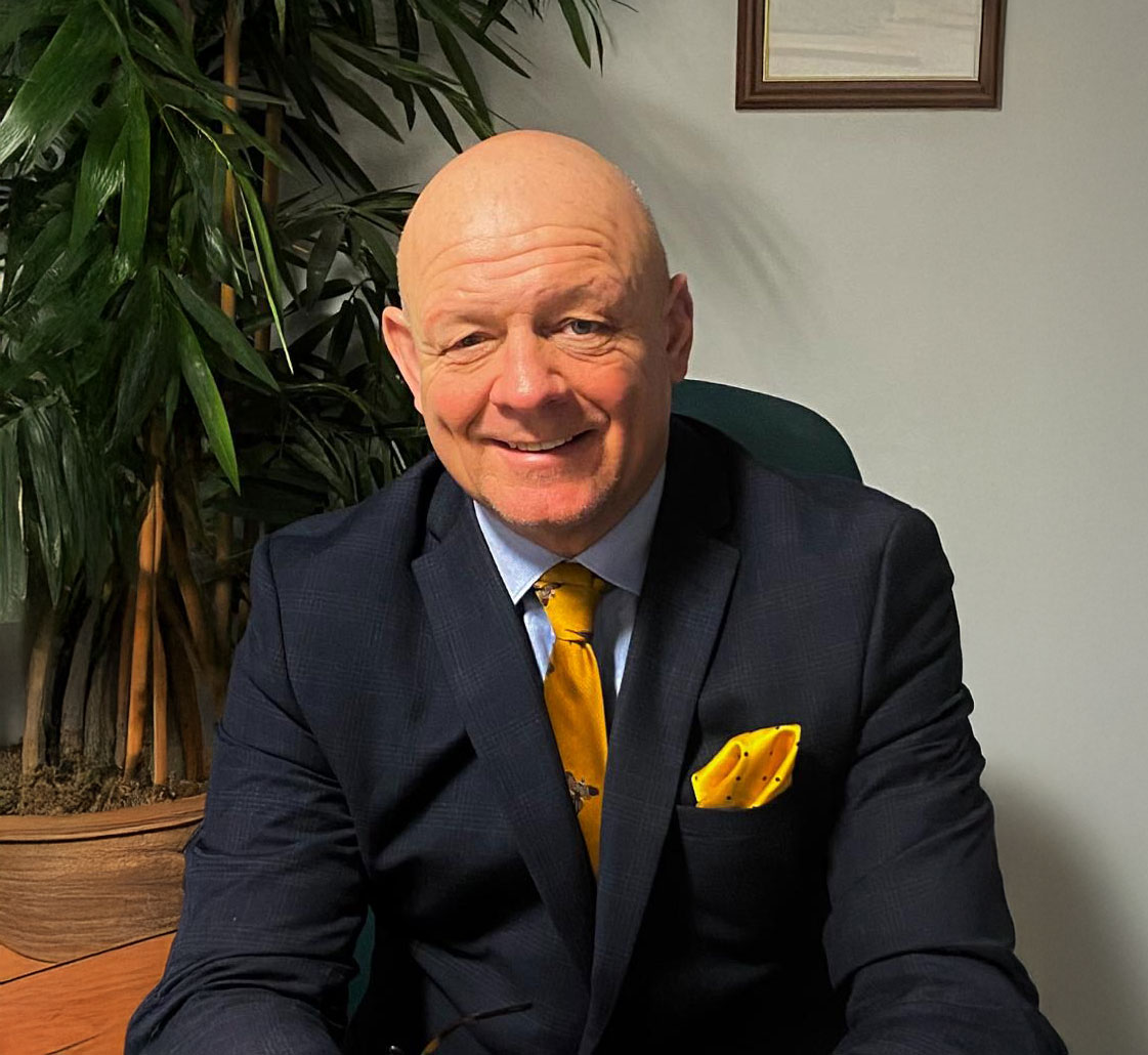 Smiling bald man in a dark suit with a yellow tie and matching pocket square seated indoors next to a large leafy plant.
