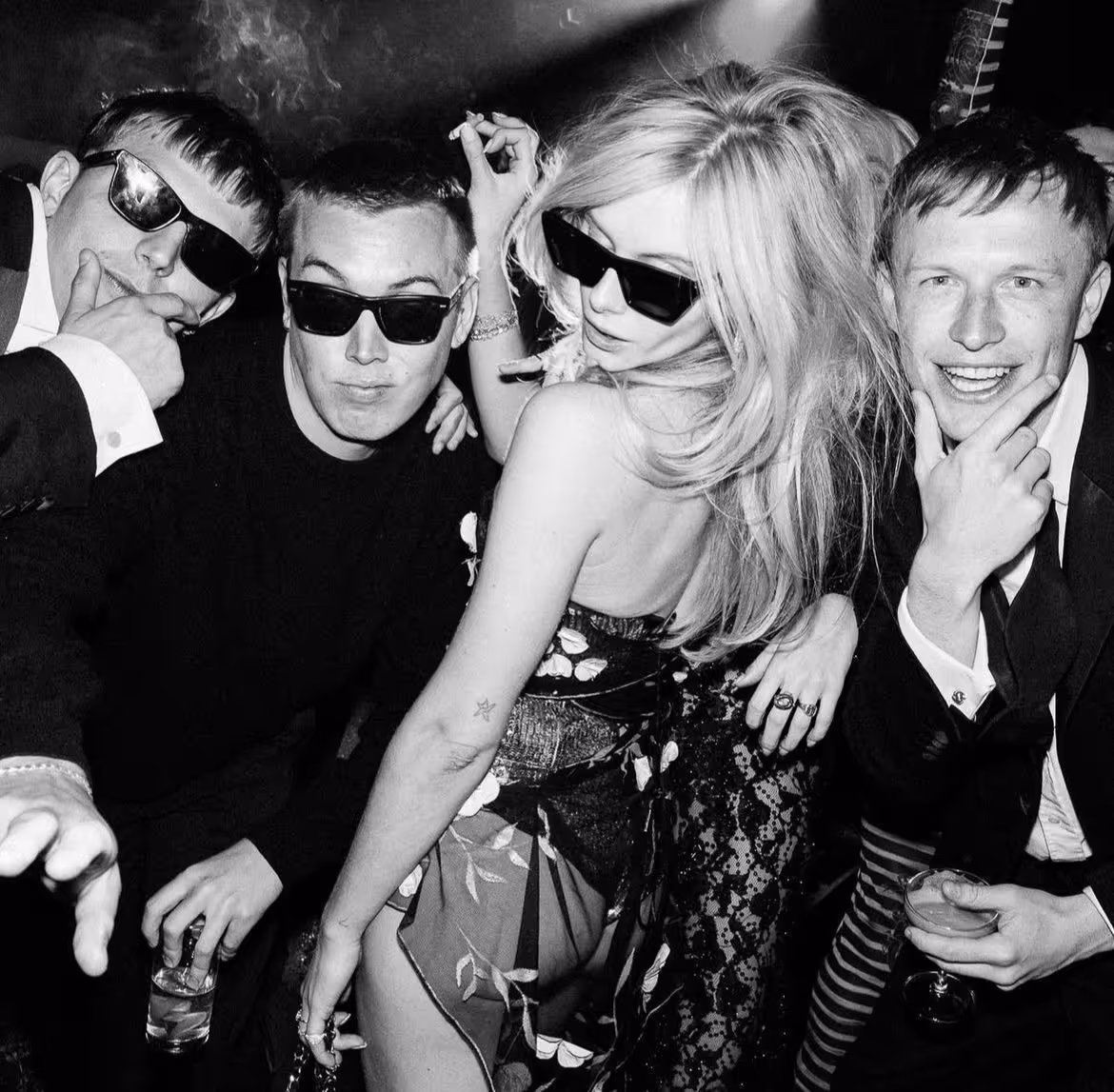 Black and white photo of four young adults wearing sunglasses, posing closely at a party, holding drinks and a cigarette.
