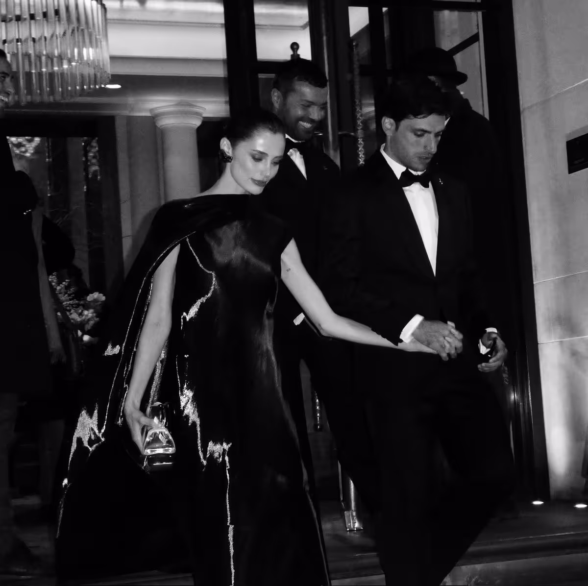 Elegant couple dressed in black formal wear holding hands as they walk down steps at a nighttime event.