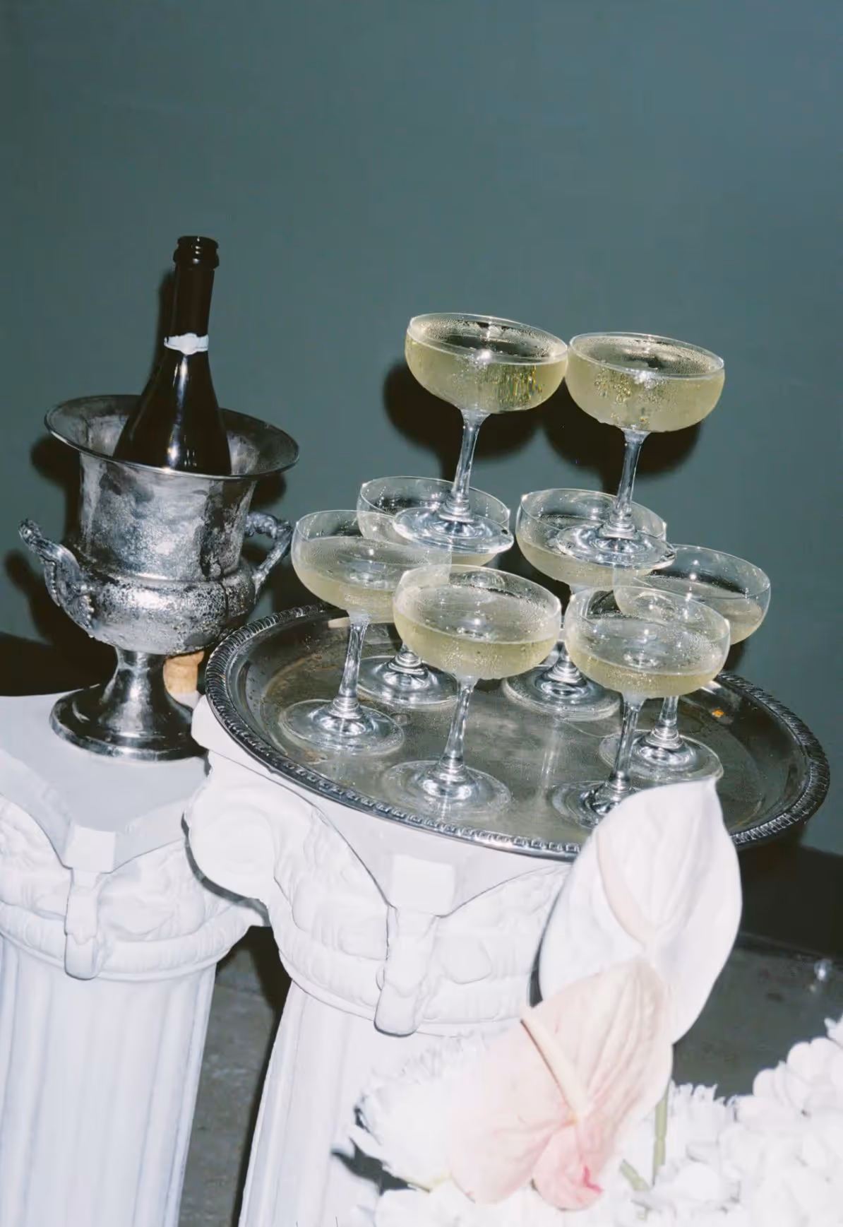 Silver tray holding seven champagne glasses filled with bubbly, next to a bottle in an ice bucket on white decorative pedestals.