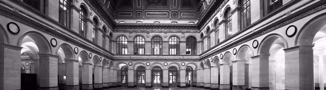 Black and white photo of a grand interior with arched windows, columns, and detailed ceiling molding.