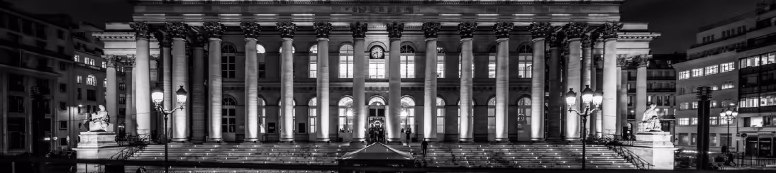 Wide nighttime view of a grand neoclassical building with tall columns and illuminated windows.