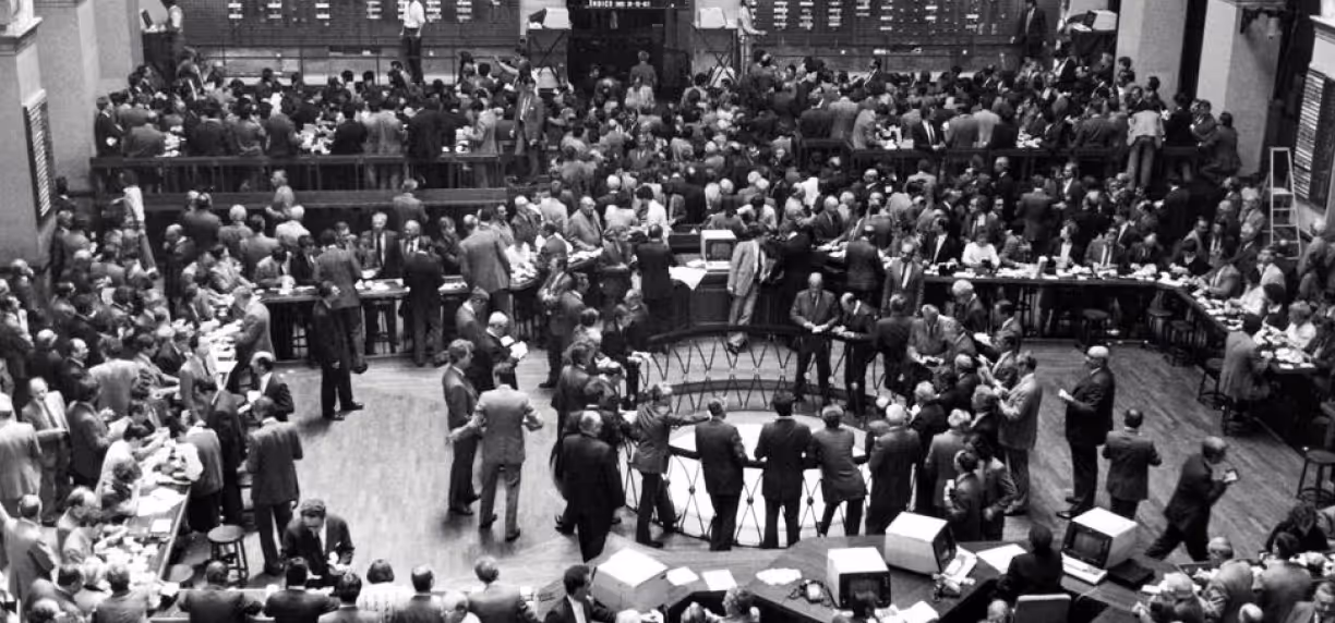 Black-and-white photo of a busy stock exchange trading floor with many men in suits gathered around desks and trading posts.