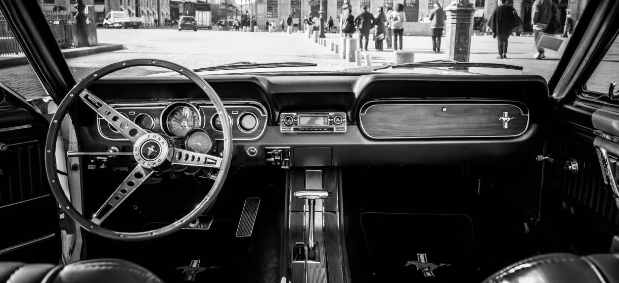 Black and white view of a classic Ford Mustang dashboard and steering wheel, with a city street visible through the windshield.