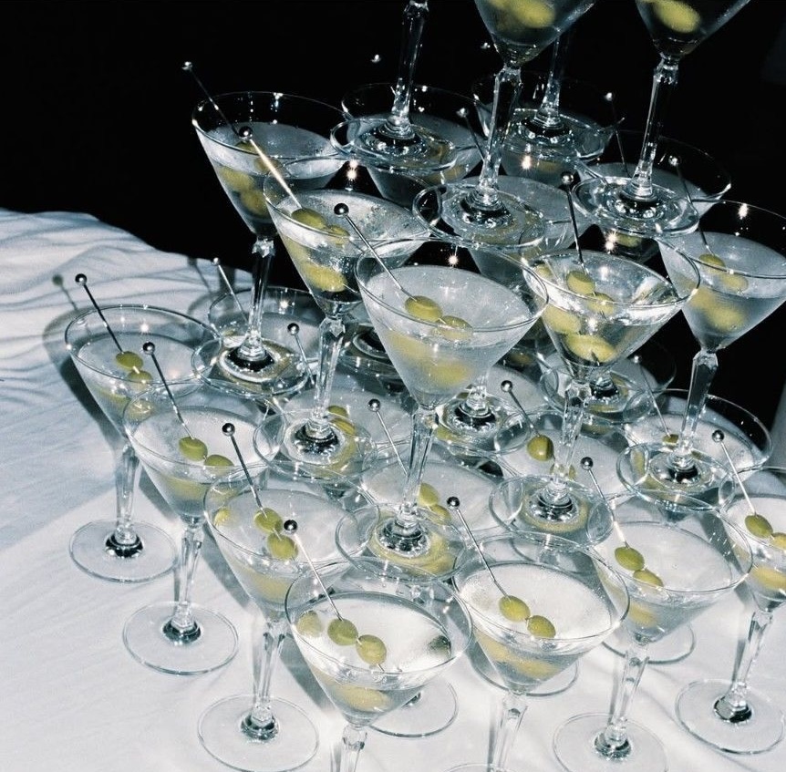 Multiple martini glasses filled with a clear liquid and garnished with green olives on cocktail picks, arranged closely on a white surface.
