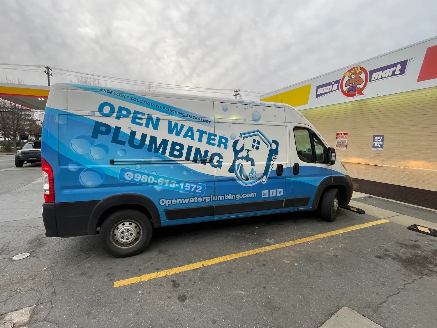 White and blue Open Water Plumbing service van parked outside a Sam's Q Mart store.