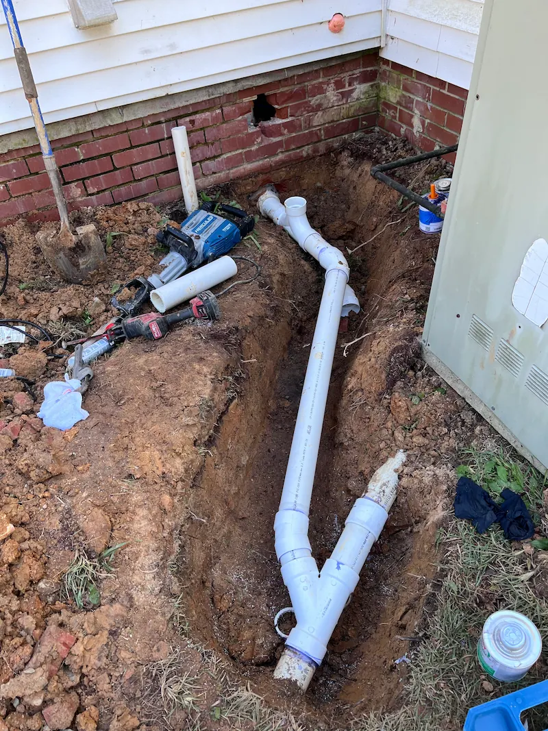 Excavated trench next to house foundation with newly installed white PVC drainage pipes and scattered plumbing tools.