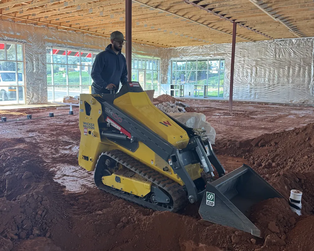 Man operating a yellow Wacker Neuson compact skid steer loader inside a building under construction with exposed wooden ceiling and dirt floor.