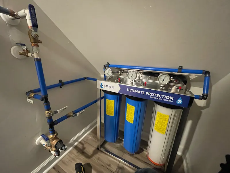Whole house water filtration system with three blue and white filter cartridges mounted on a metal frame with pressure gauges and blue piping.
