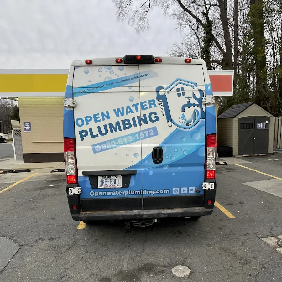 Rear view of a white and blue plumbing service van with text 'Open Water Plumbing' and contact number parked at a gas station.