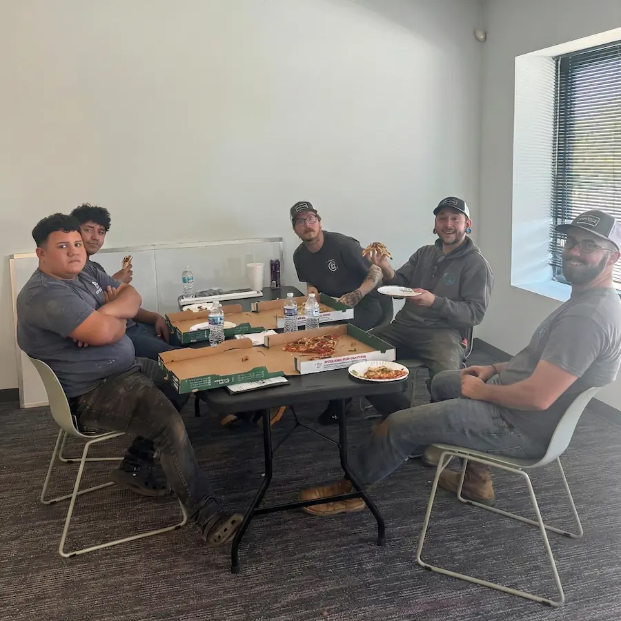 Five men sitting around a table eating pizza, with pizza boxes and water bottles on the table in a bright room.