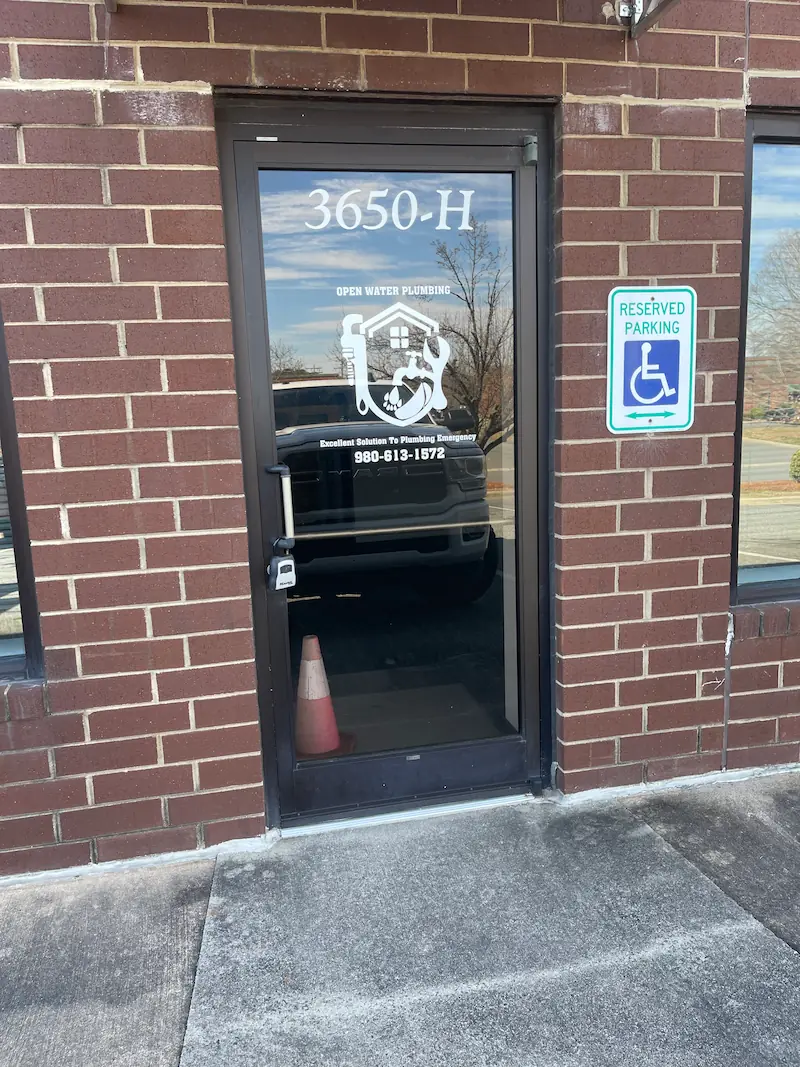 Glass door of a brick building labeled 3650-H with Open Water Plumbing logo and phone number, next to a reserved parking sign for handicapped.