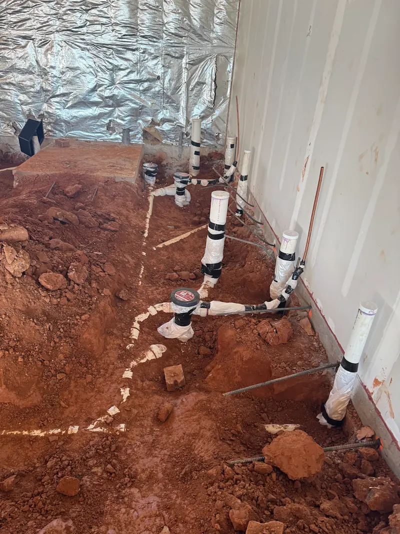 Indoor construction site showing exposed plumbing pipes wrapped in protective material emerging from red soil floor inside an unfinished room.