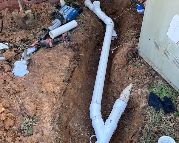 Trench next to a brick house with newly installed white PVC drainage pipes and plumbing tools scattered around.