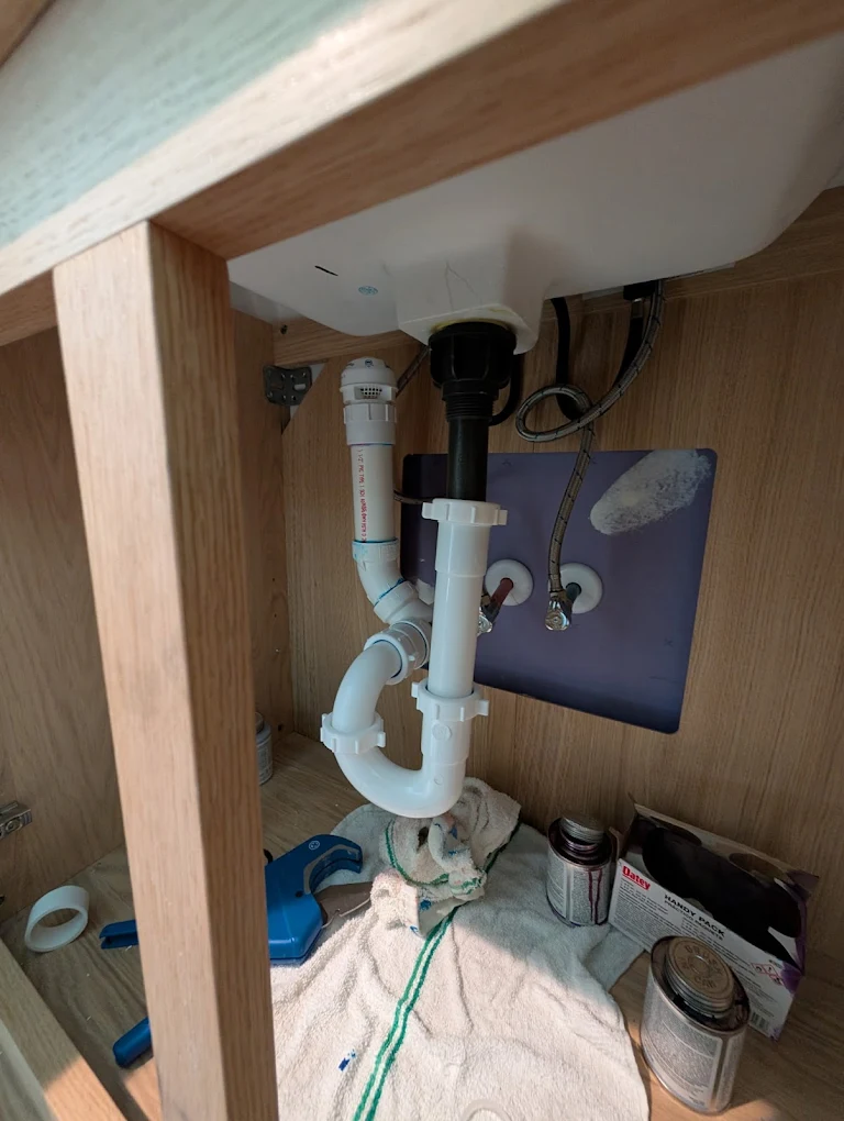 Trench dug beside a brick house with white PVC plumbing pipes being installed and power tools lying nearby.