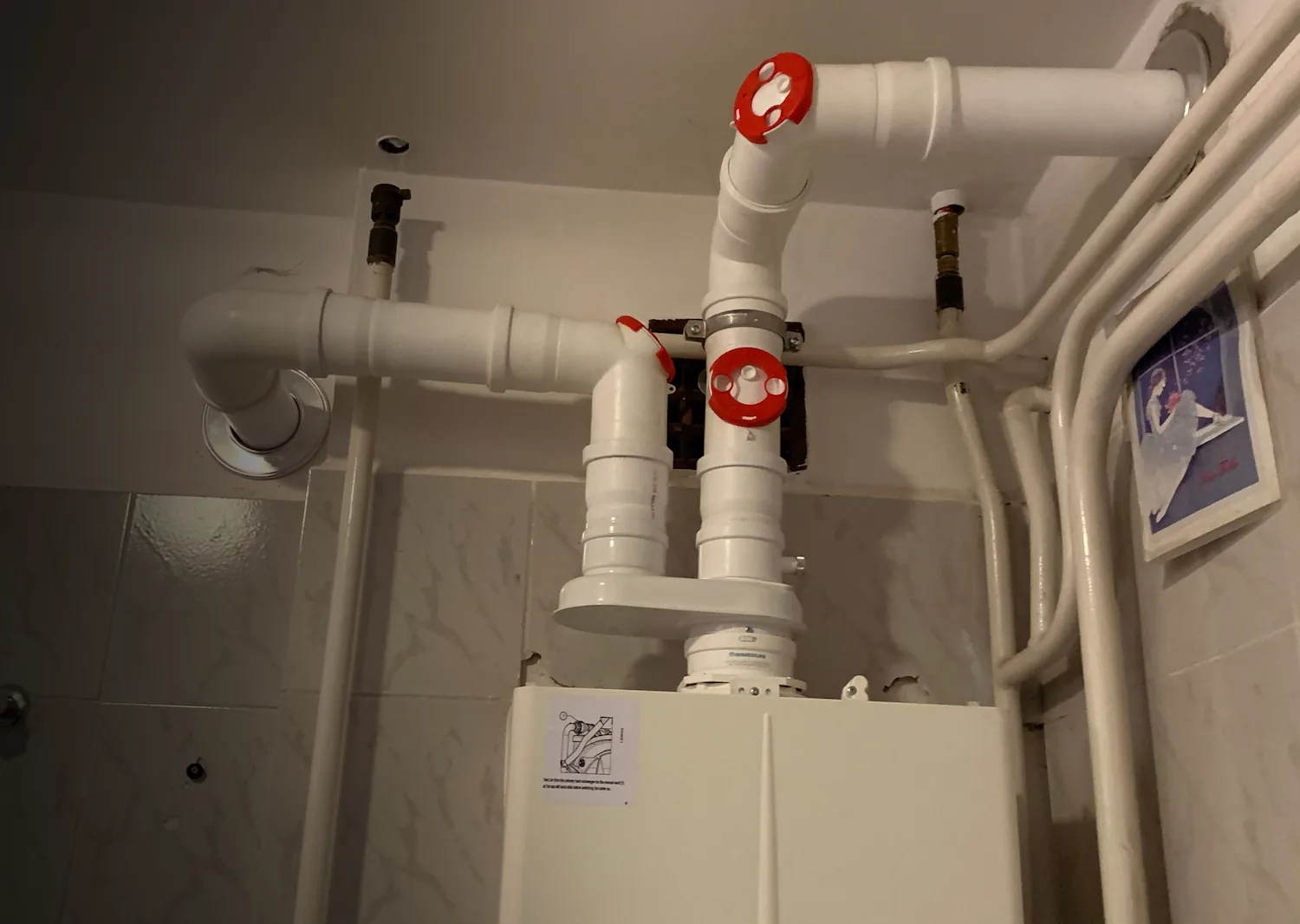 White pipes with red valve handles connected to a boiler or heating unit on tiled wall.