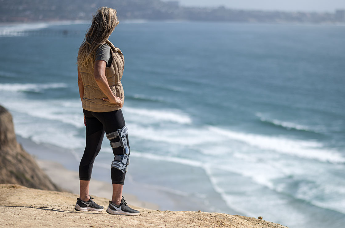 Women standing on cliff above beach wearing a DonJoy OA Nano in her right leg.
