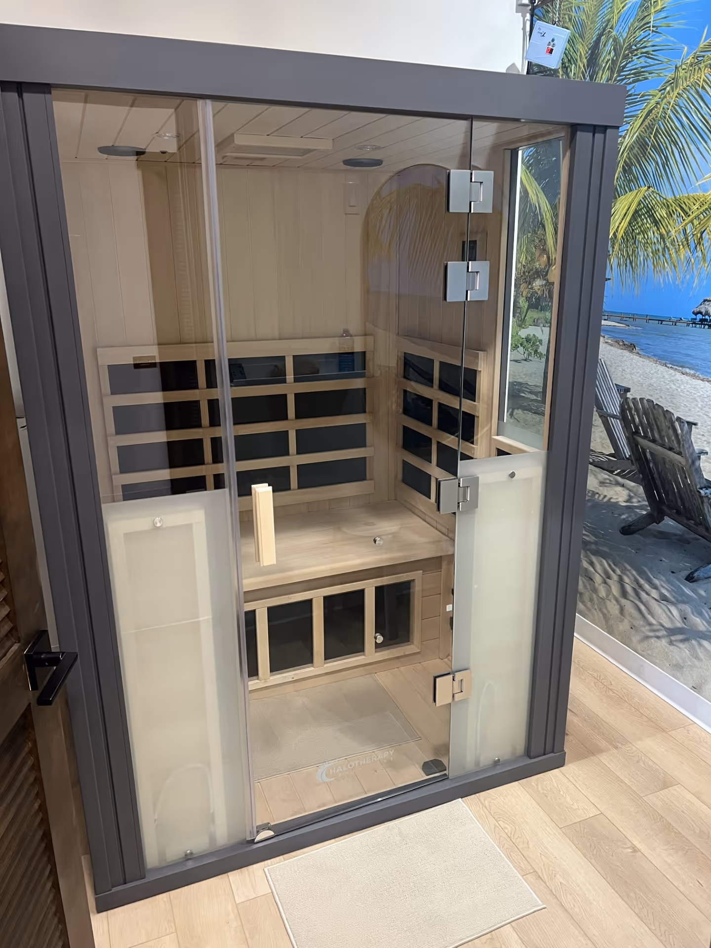 Indoor wooden sauna with a glass door and bench, next to a wall mural of a beach scene with palm trees and chairs.