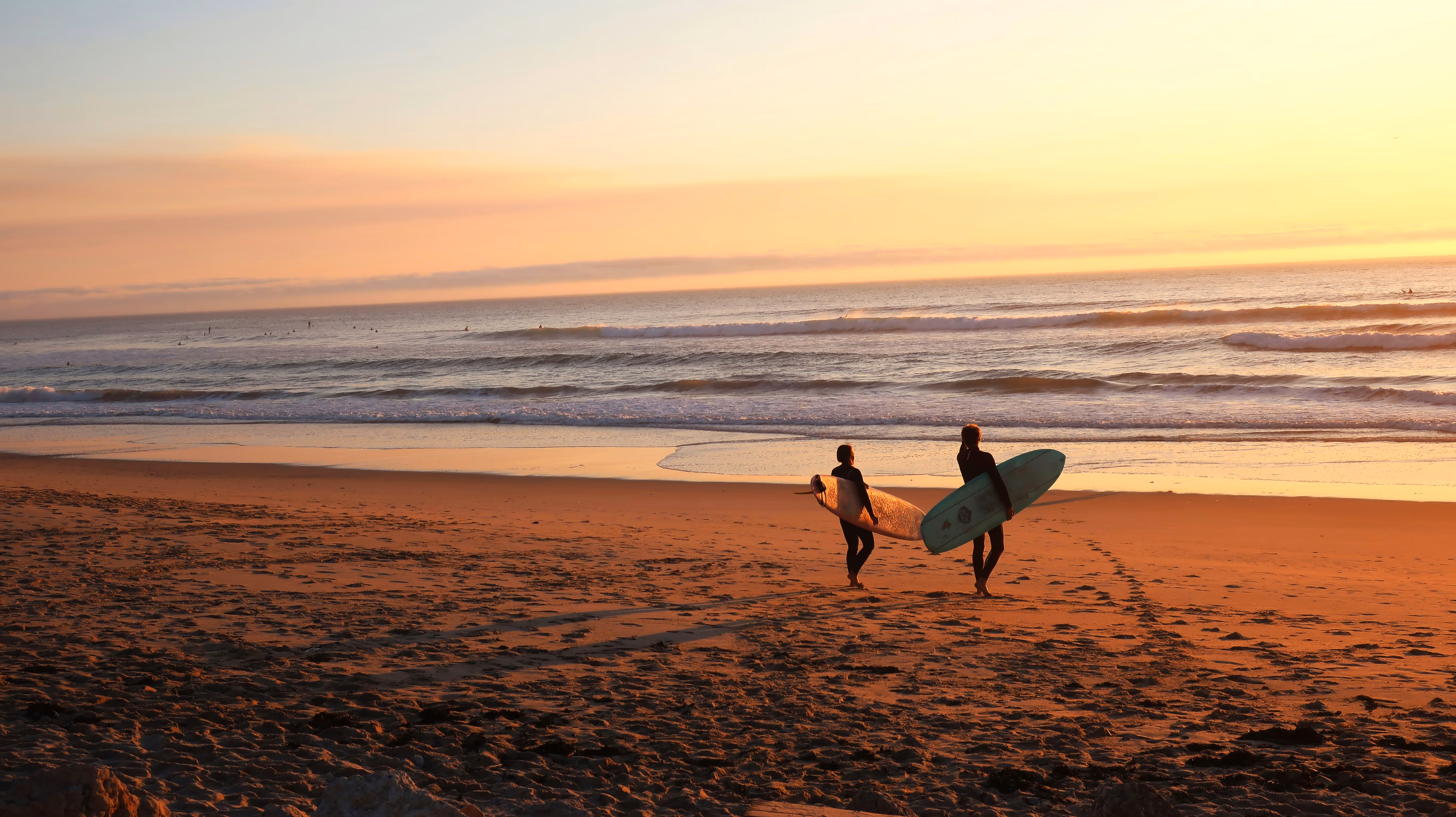 Two surfers carrying surfboards walking on a sandy beach at sunset toward the ocean waves.