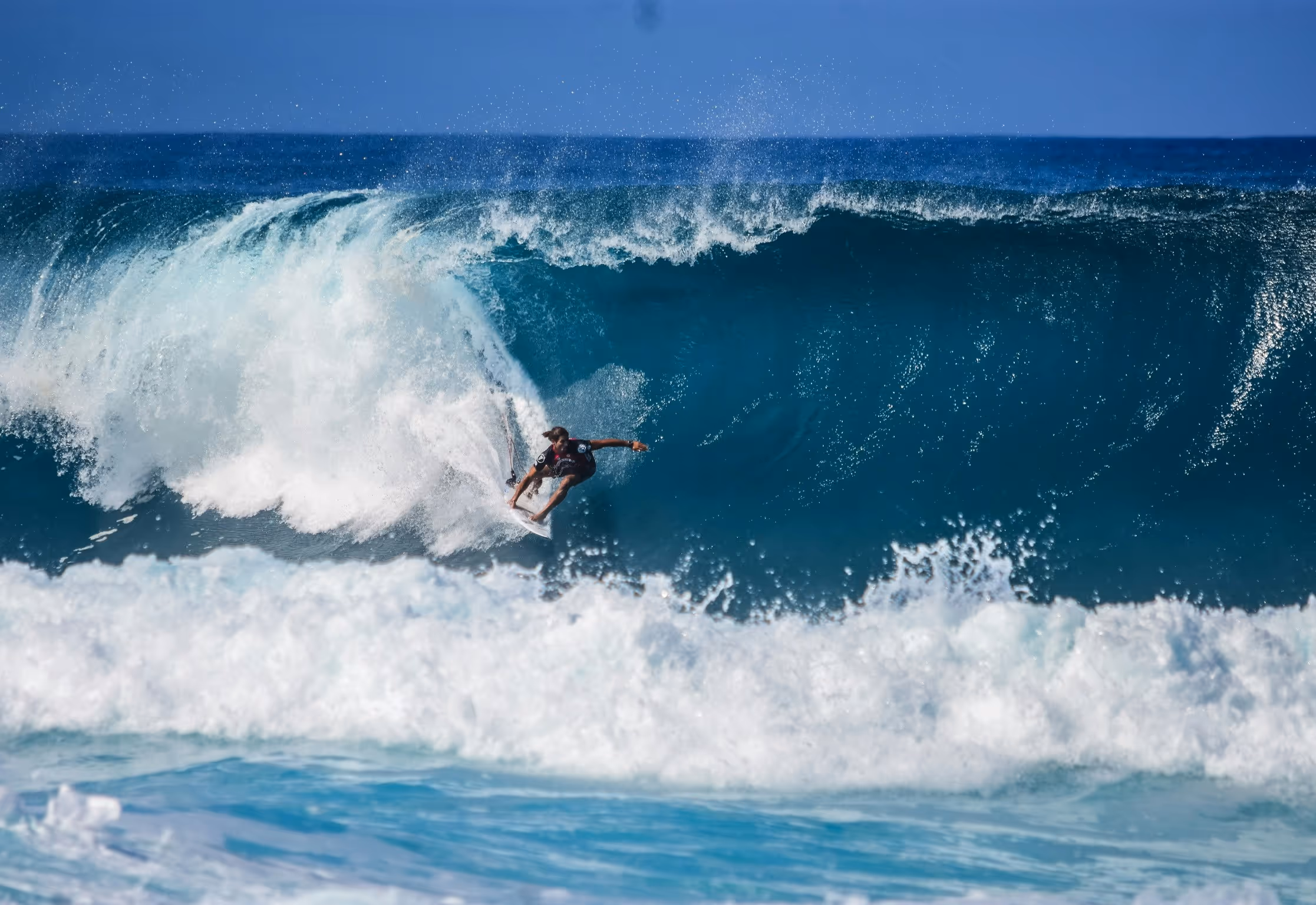 Surfer carving through a large blue ocean wave with white spray.