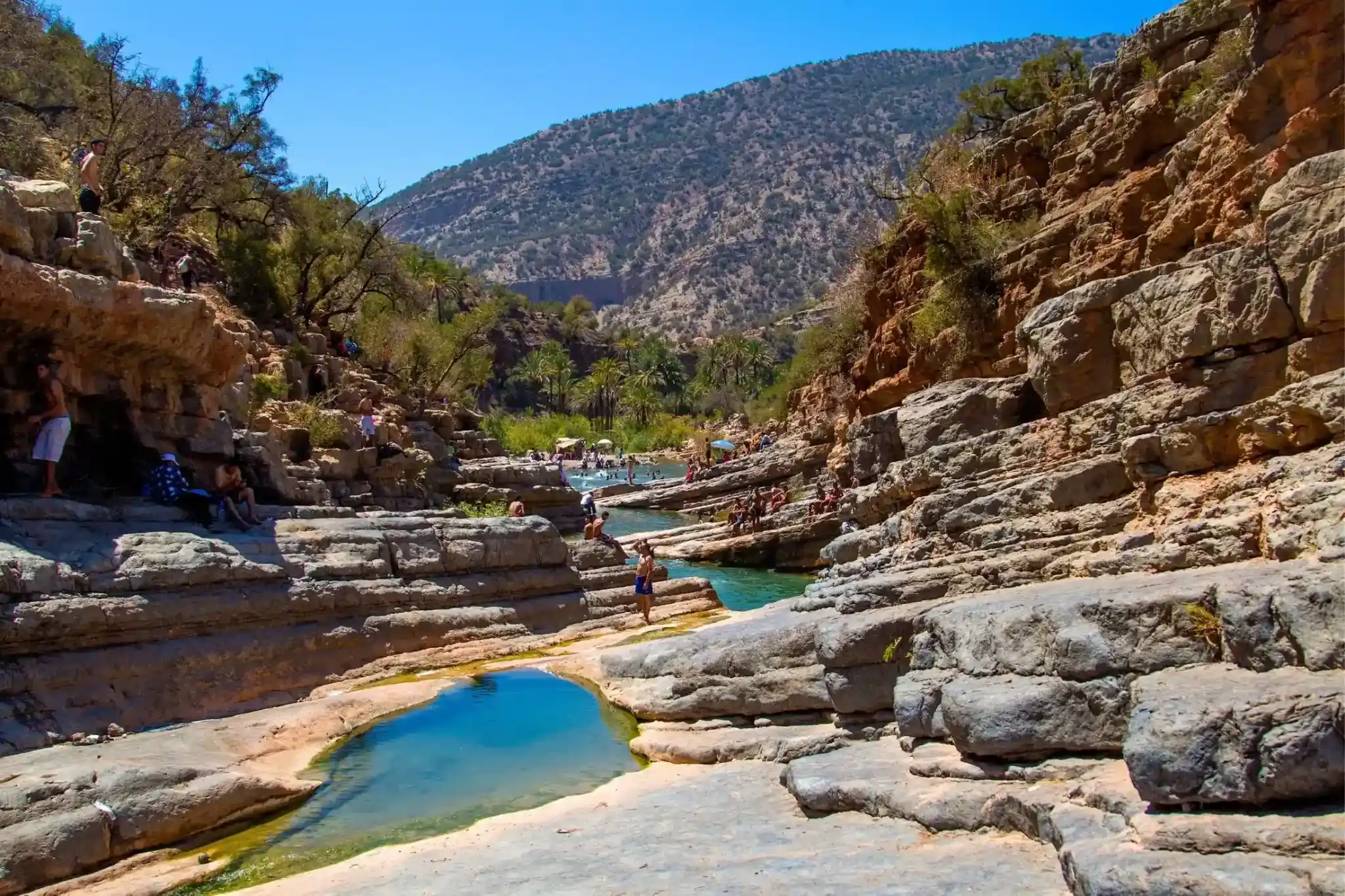 Rocky canyon with turquoise water pools and people enjoying the sunny day surrounded by vegetation and mountains.