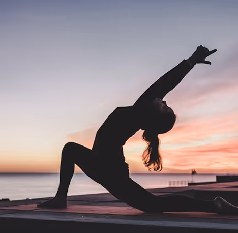 Silhouette of a person performing a low lunge yoga pose at sunset by the water.