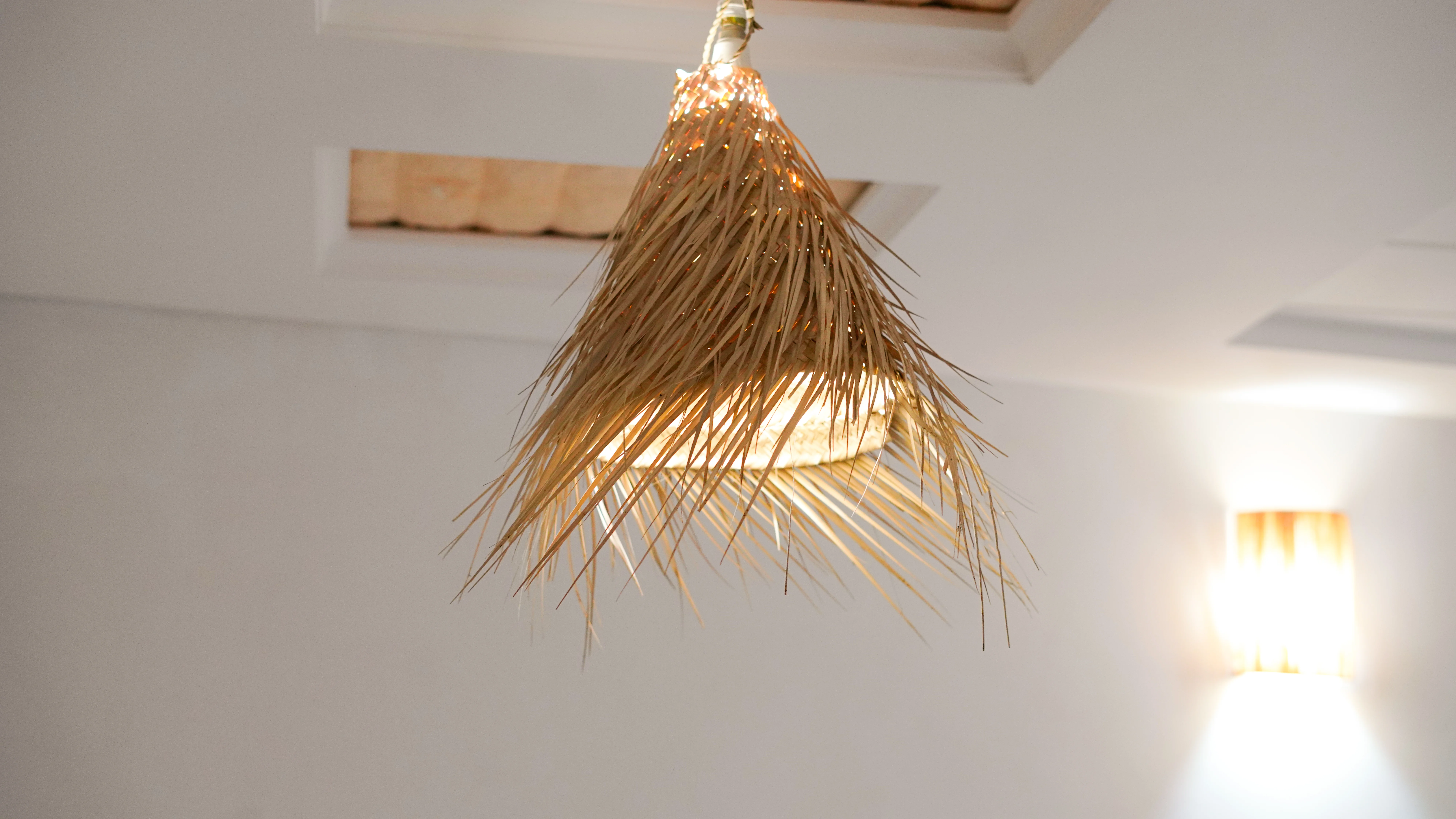 Hanging ceiling light fixture with a lampshade made of woven natural straw material, glowing warmly in a bright room.