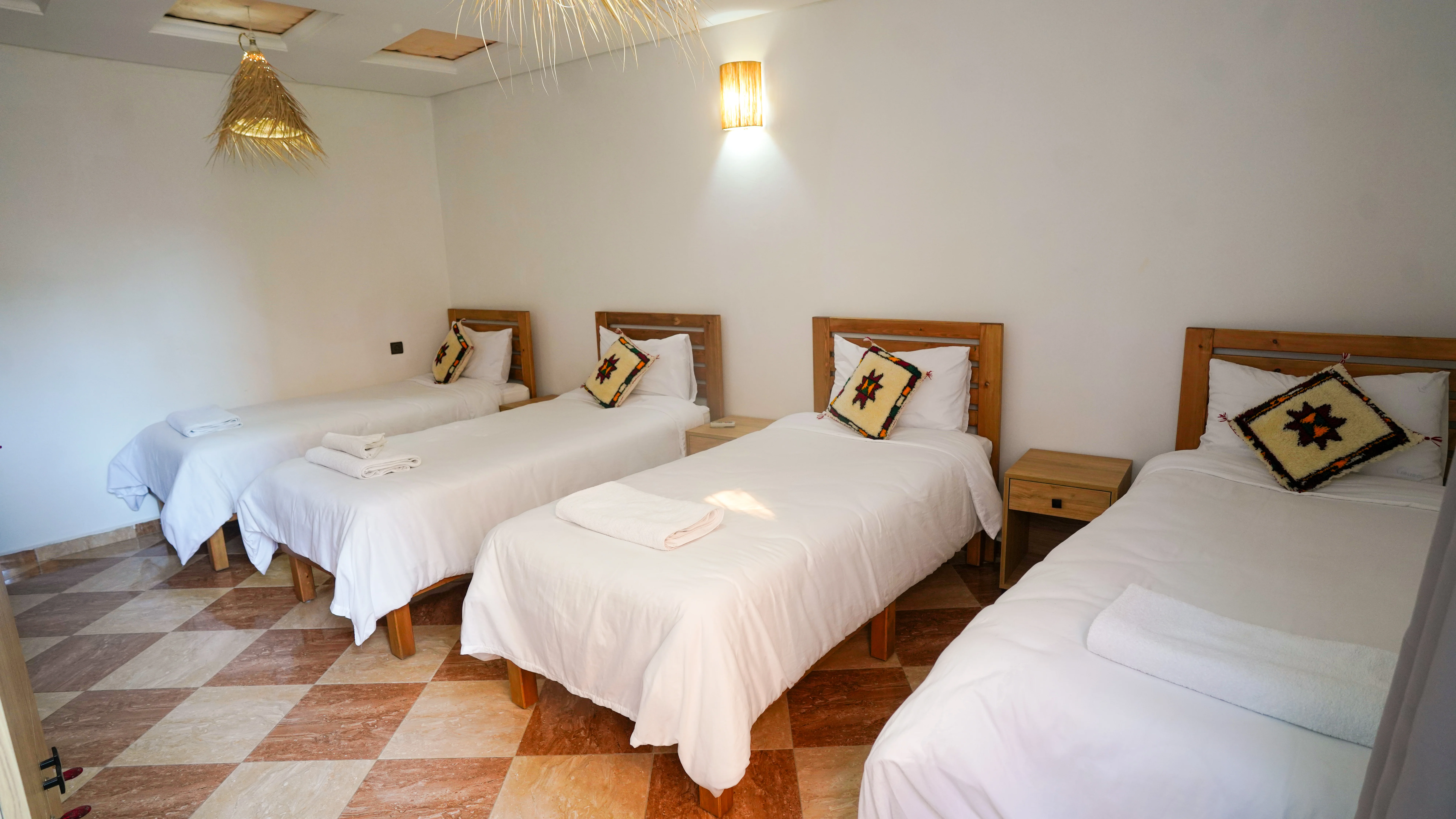 Four single beds with white linens and colorful patterned pillows in a room with a checkered brown and beige tile floor.