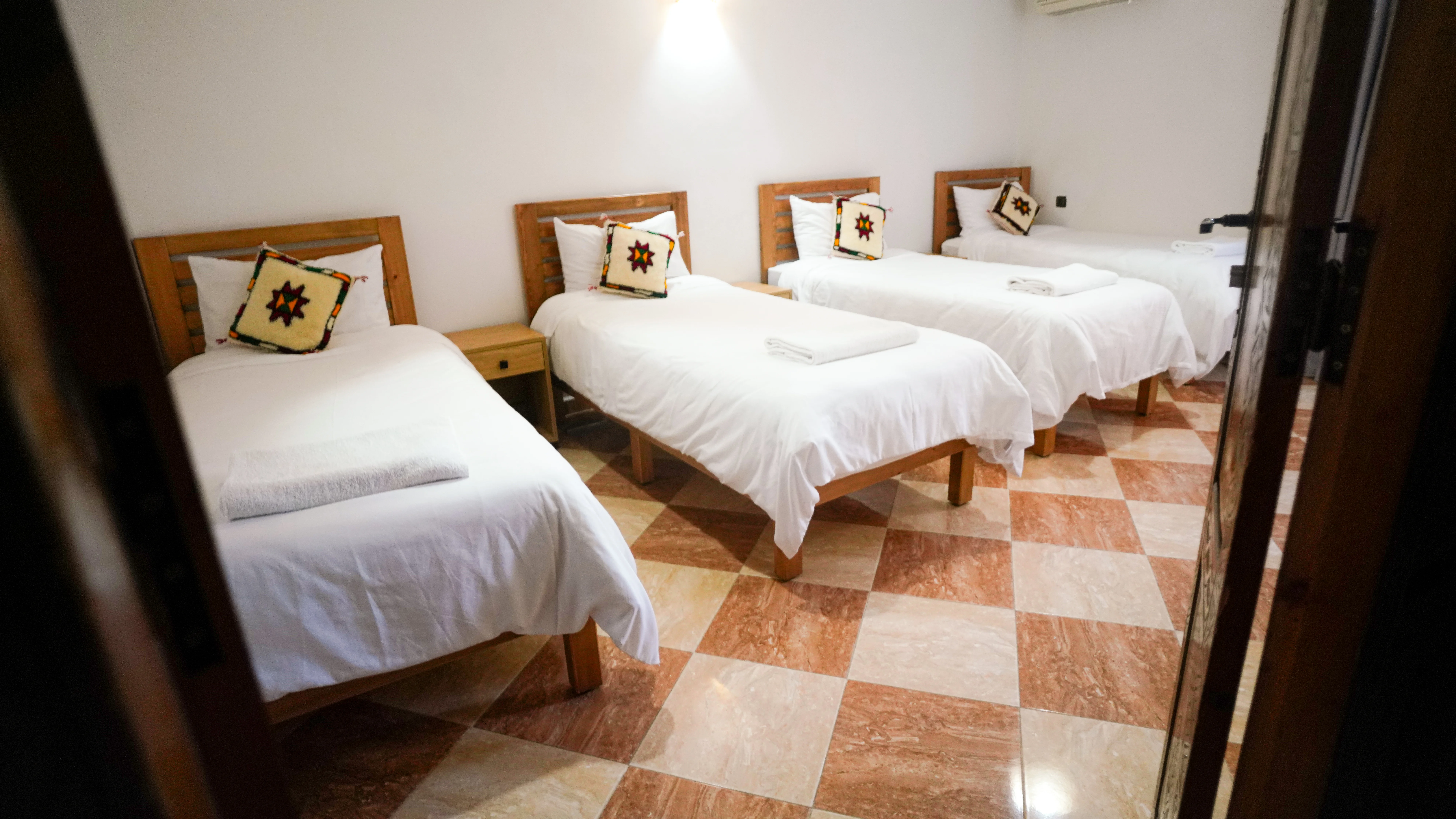 Four single beds with white linens and decorative pillows in a room with checkered tile floor.