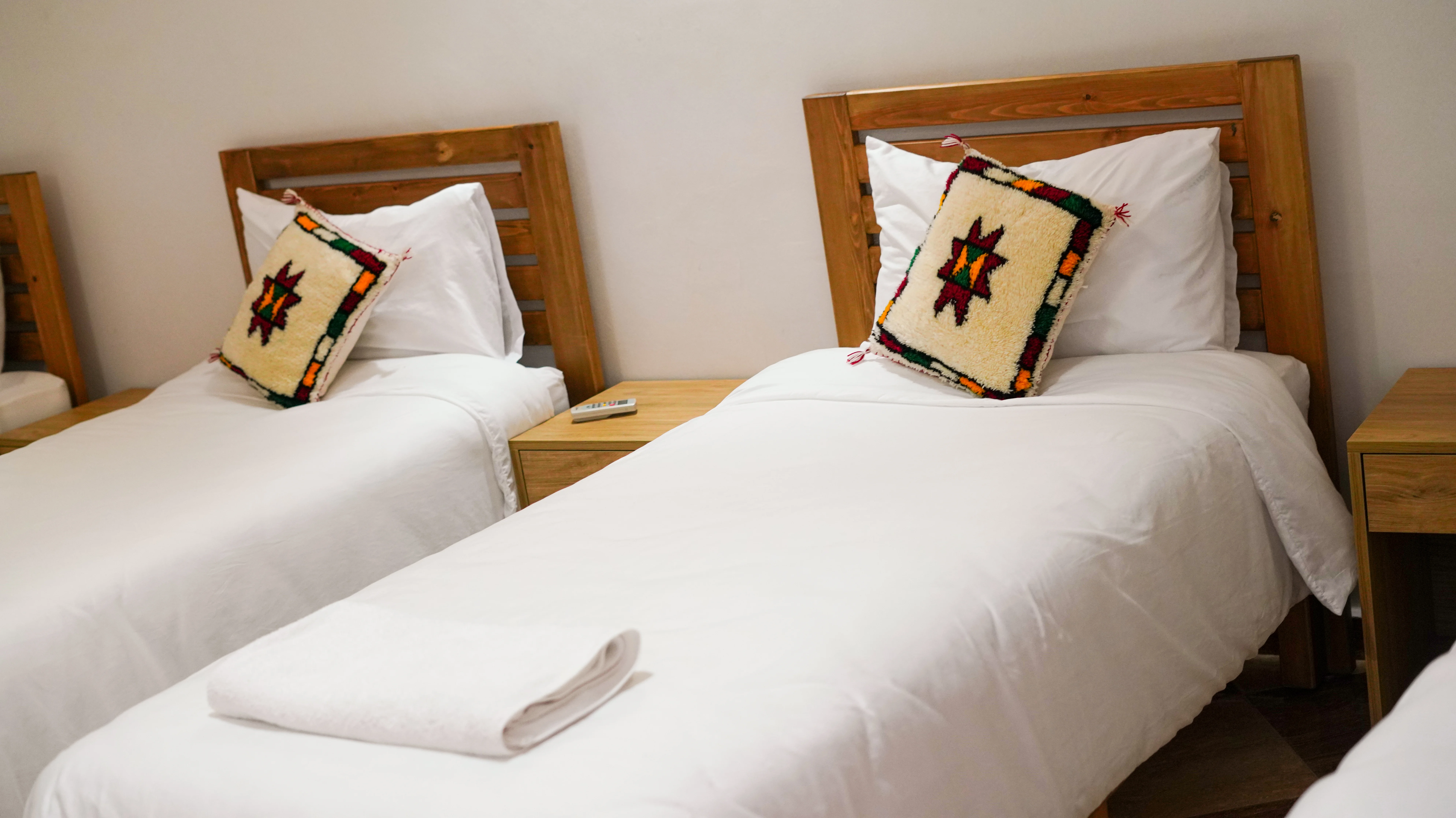 Two single beds with wooden headboards, white bedding, patterned decorative pillows, and a folded towel on one bed.