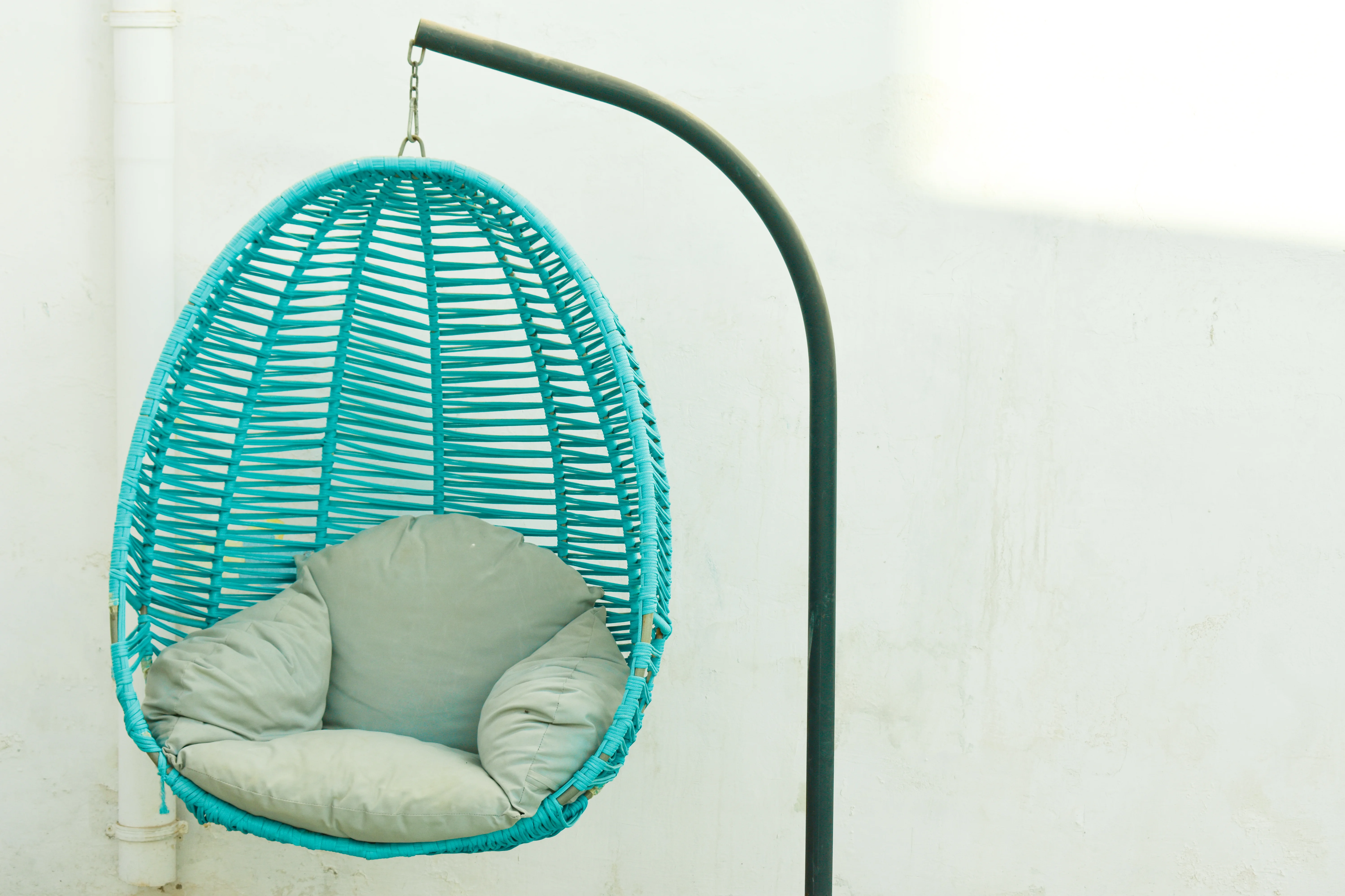 Blue woven hanging chair with gray cushions suspended from a black metal stand against a white wall.