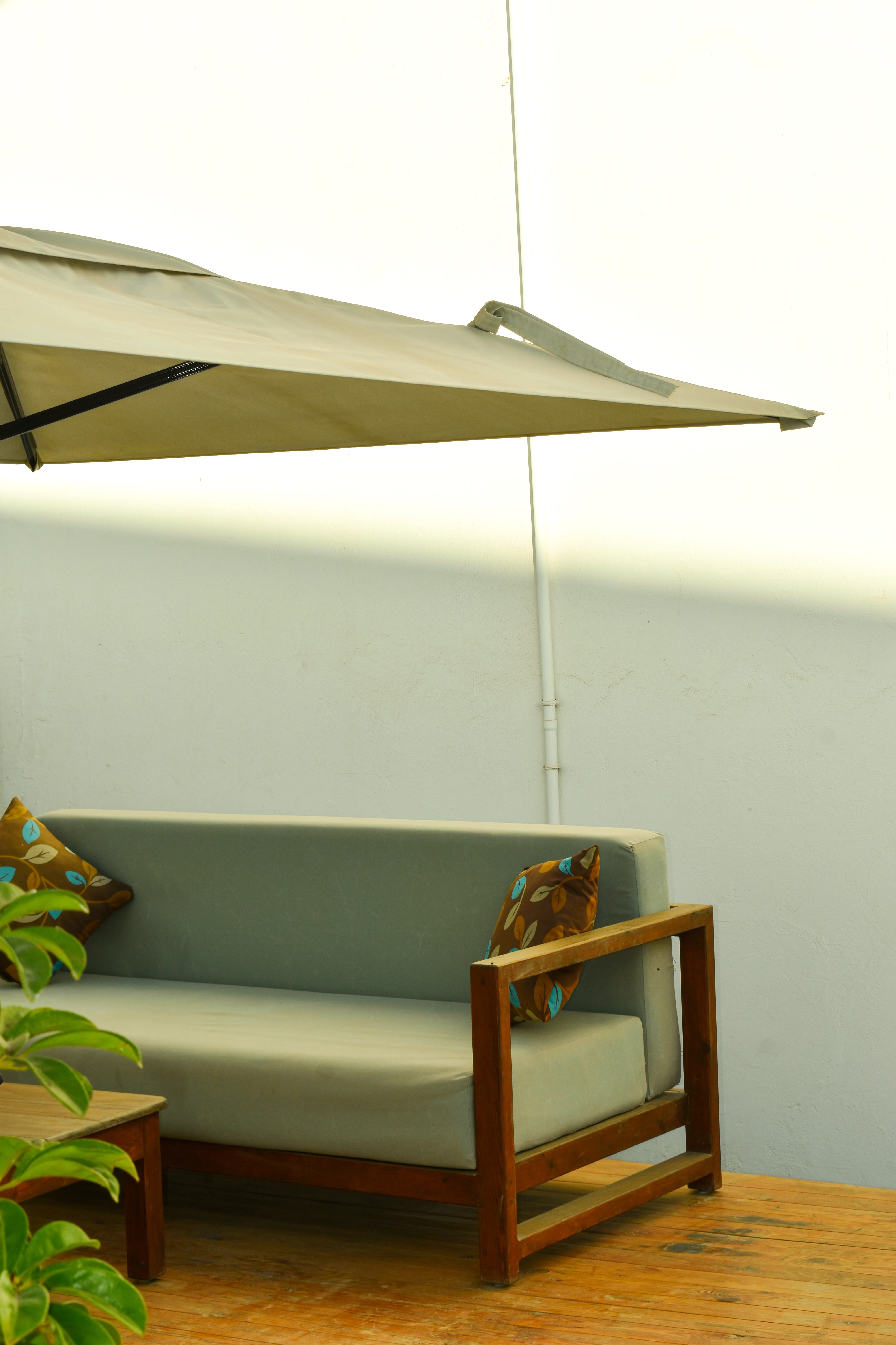 Outdoor wooden-framed sofa with green cushions and leaf-patterned pillows under a large beige umbrella on a wooden deck.