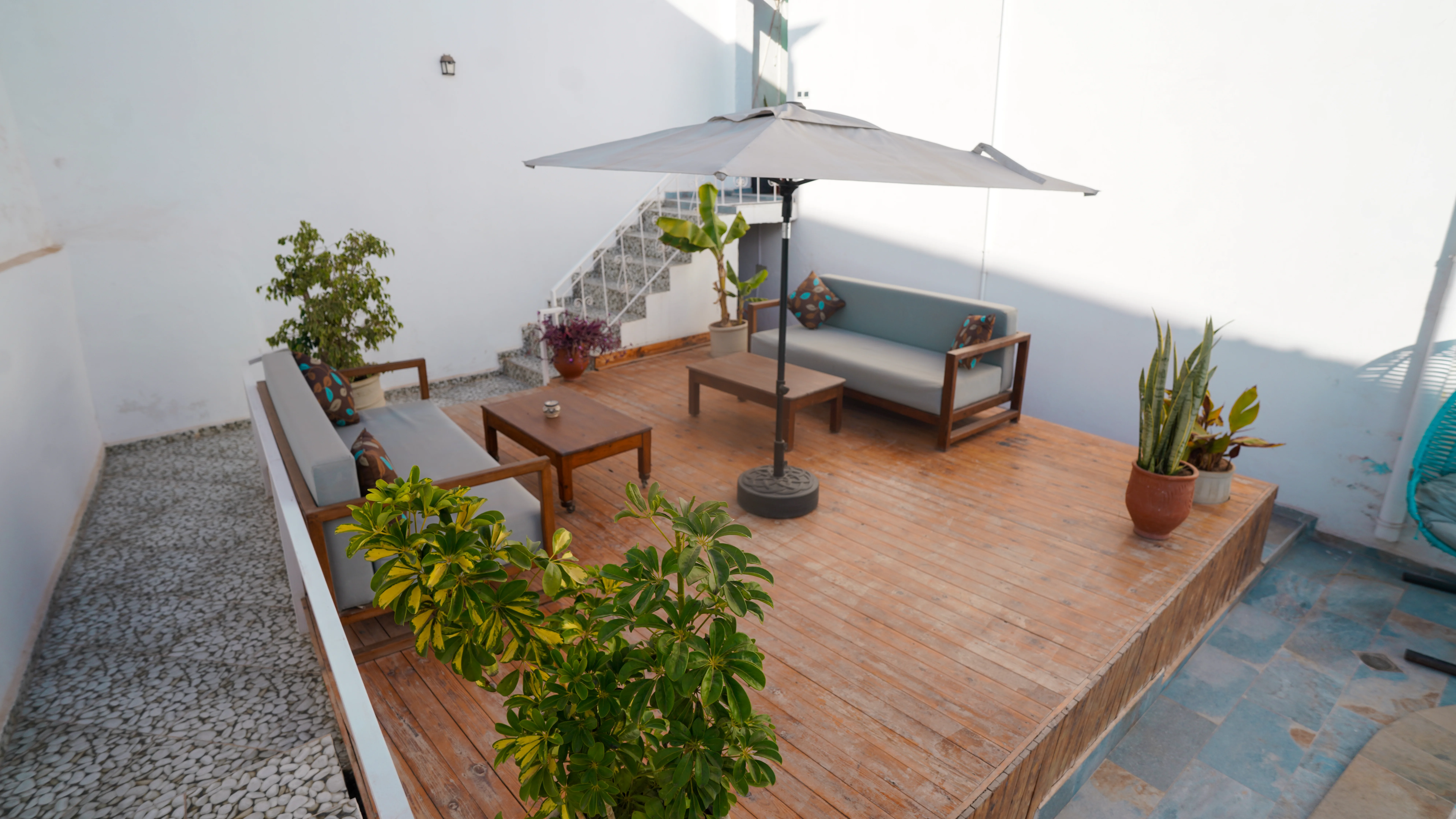 Outdoor wooden deck with two gray cushioned sofas, a central umbrella, wooden coffee tables, and several potted plants.