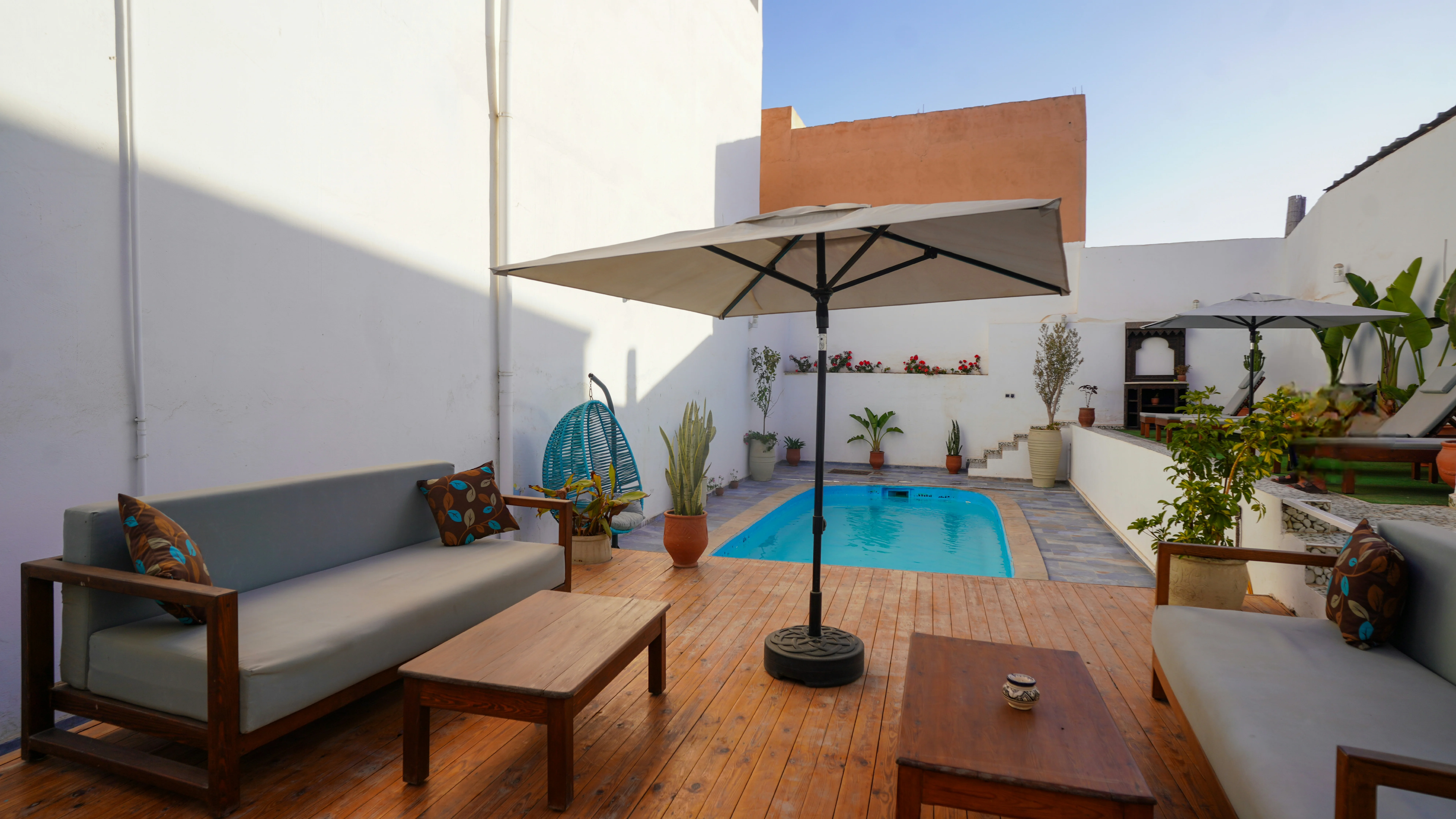 Modern outdoor patio with wooden deck, gray cushioned sofas, coffee tables, large umbrella, small blue swimming pool, and potted plants.