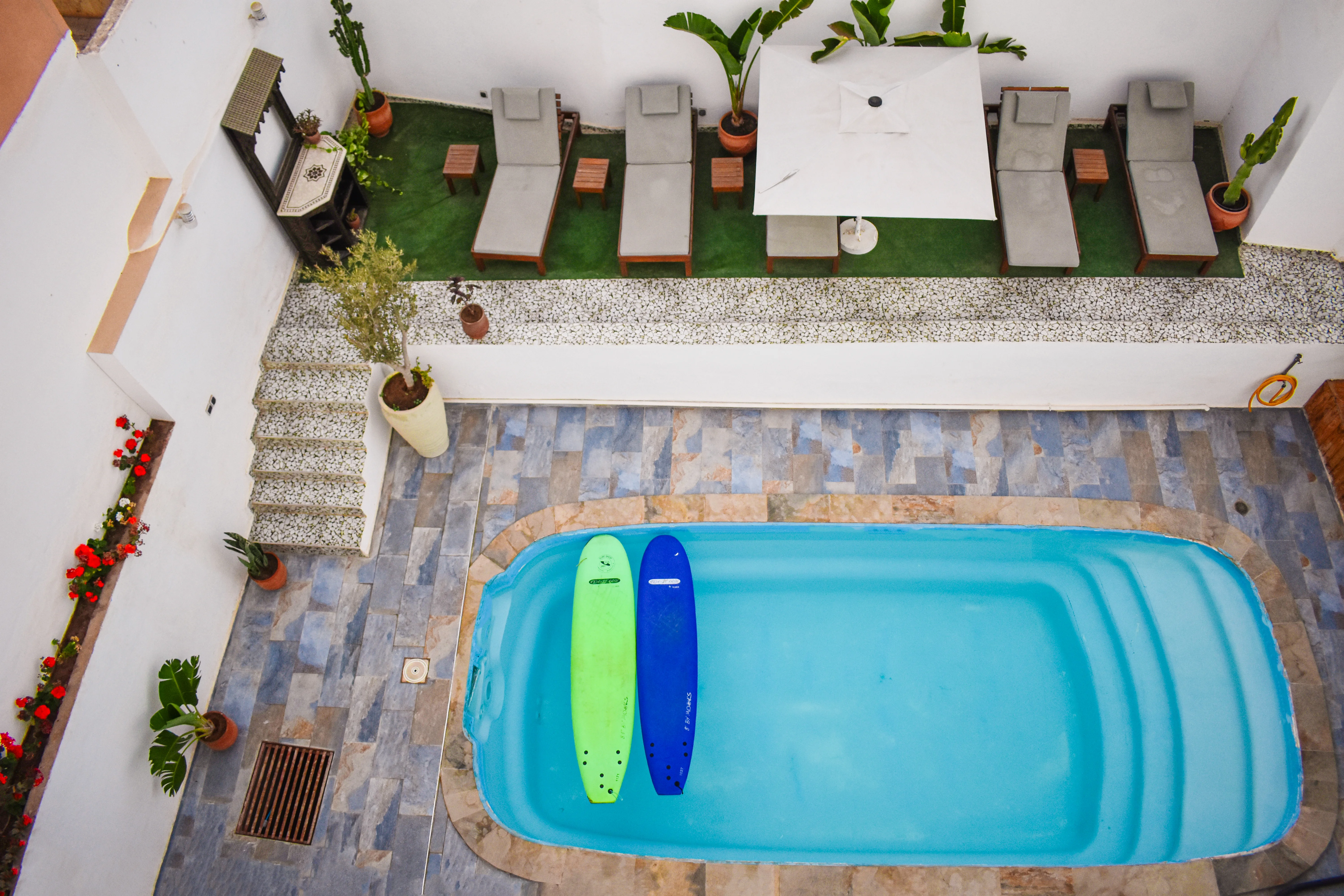 Small outdoor swimming pool with two surfboards floating, surrounded by loungers, potted plants, and tiled flooring.