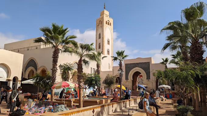 Bustling market scene with palm trees, shoppers, and a tall mosque tower under a blue sky.