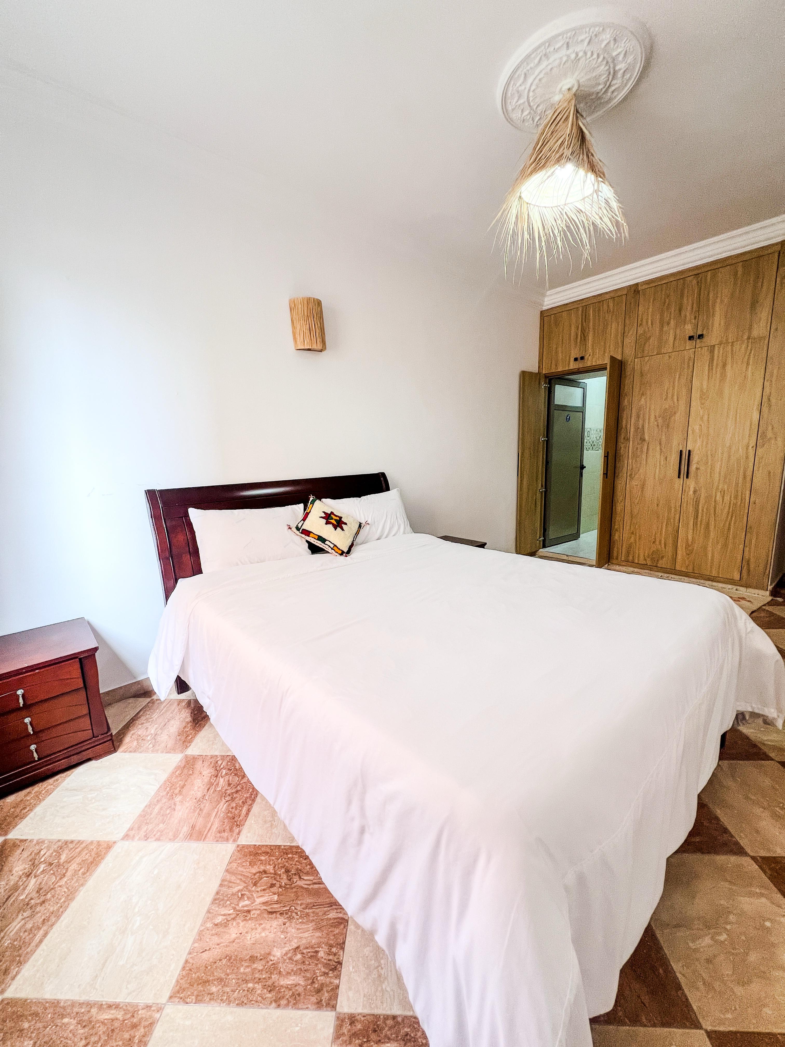 Bedroom with a large bed covered in white linens, a wooden headboard, a colorful accent pillow, wooden nightstand, checkered tile floor, and a ceiling light with a natural fiber shade.