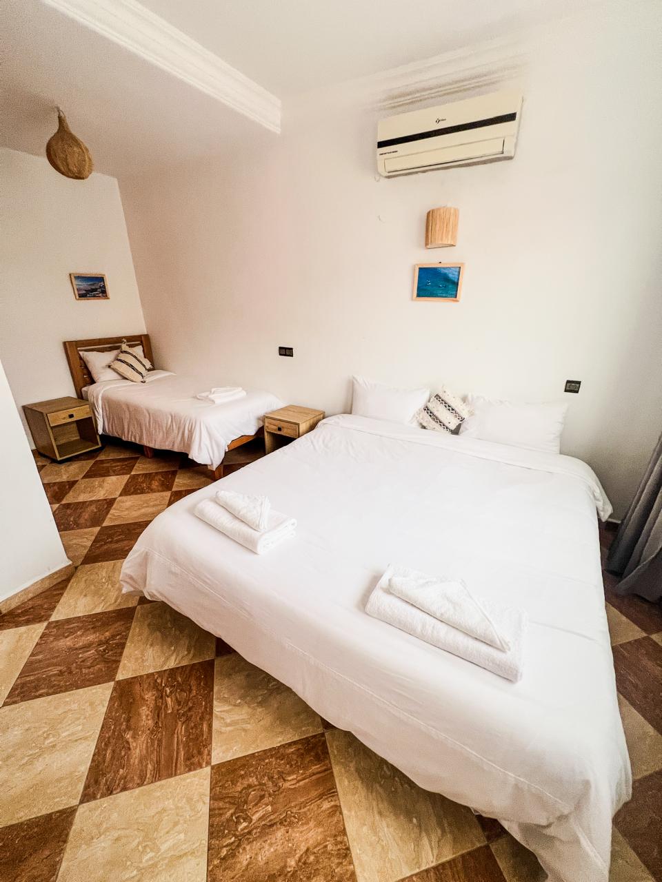 Bedroom with a large bed and a single bed, both with white linens and folded towels on them, checkered brown and beige floor, air conditioner on wall.