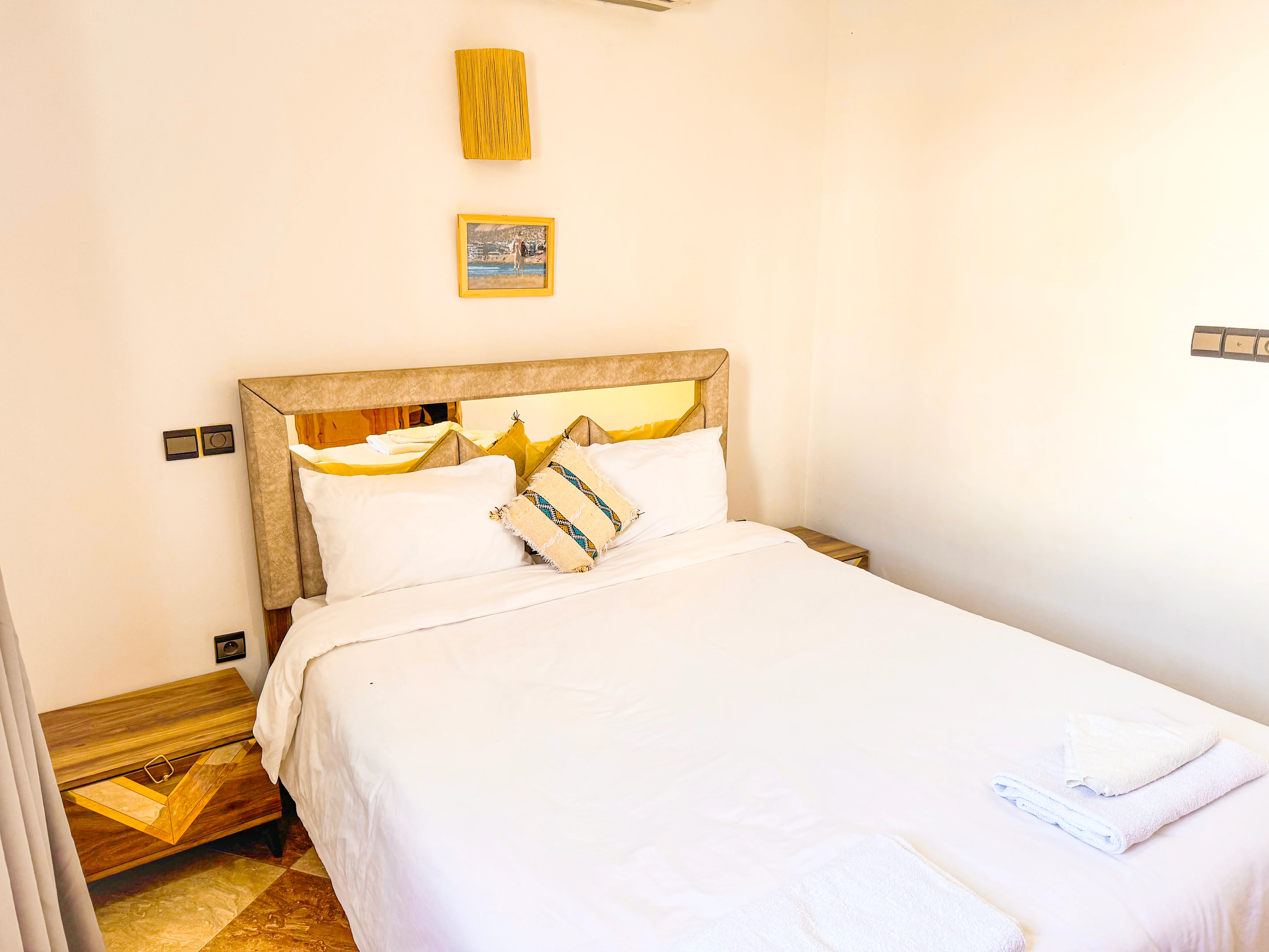 Bright bedroom with a neatly made double bed, white linens, decorative pillows, wooden nightstands, and a wall lamp.
