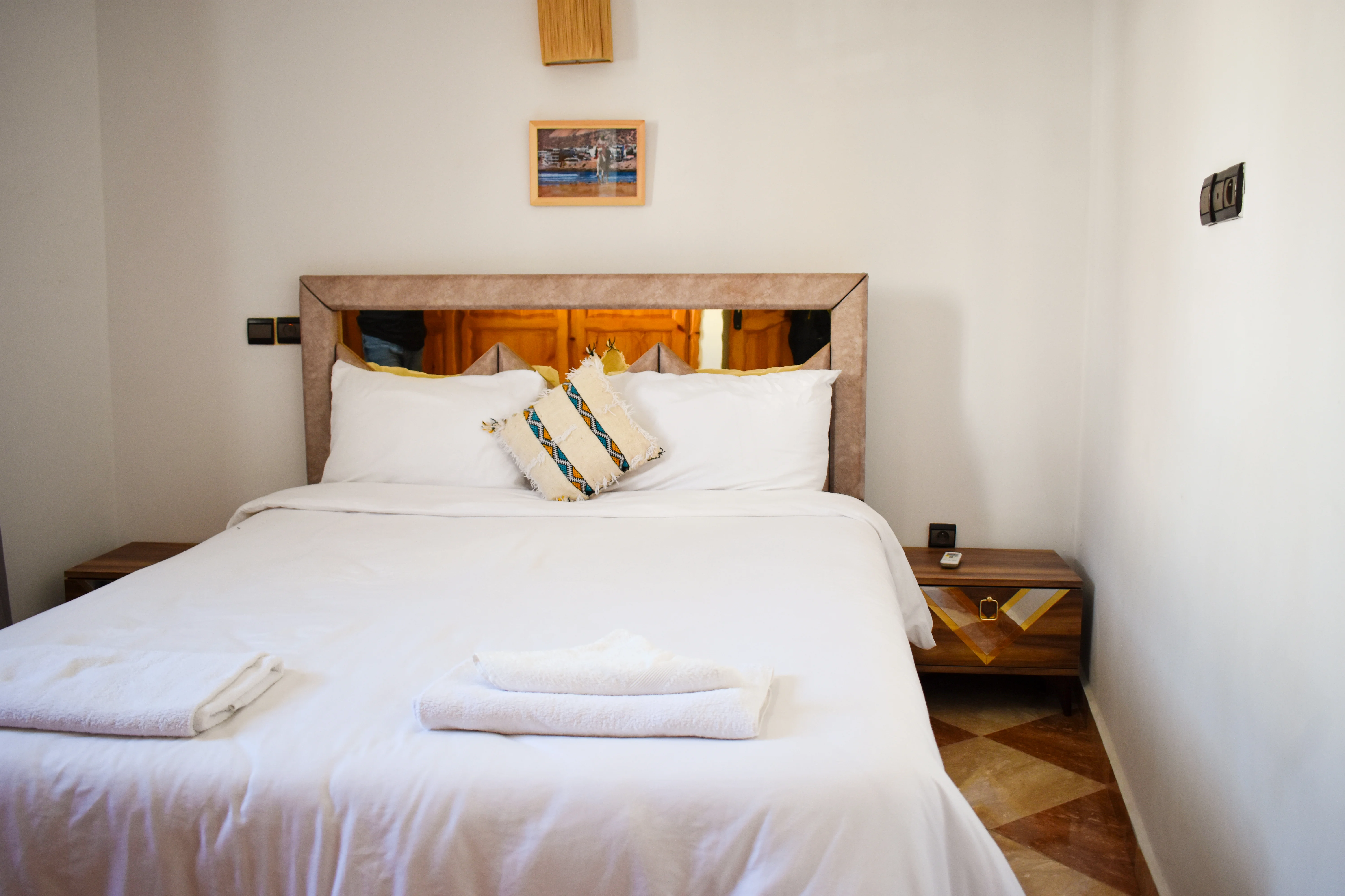 Bed with white linens, two white pillows, a decorative pillow, and two folded towels at the foot, flanked by wooden nightstands against a plain white wall.